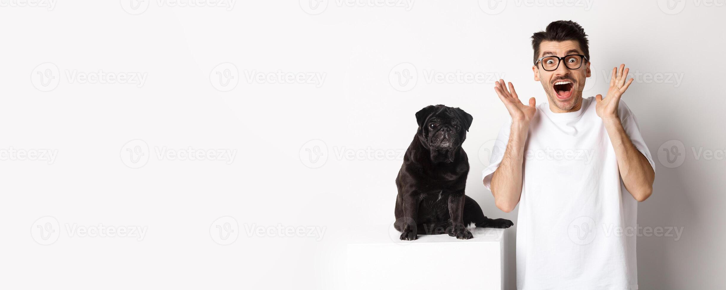Ecstatic young man looking with excitement and rejoice, standing near cute black pug, staring at camera happy, standing over white background photo