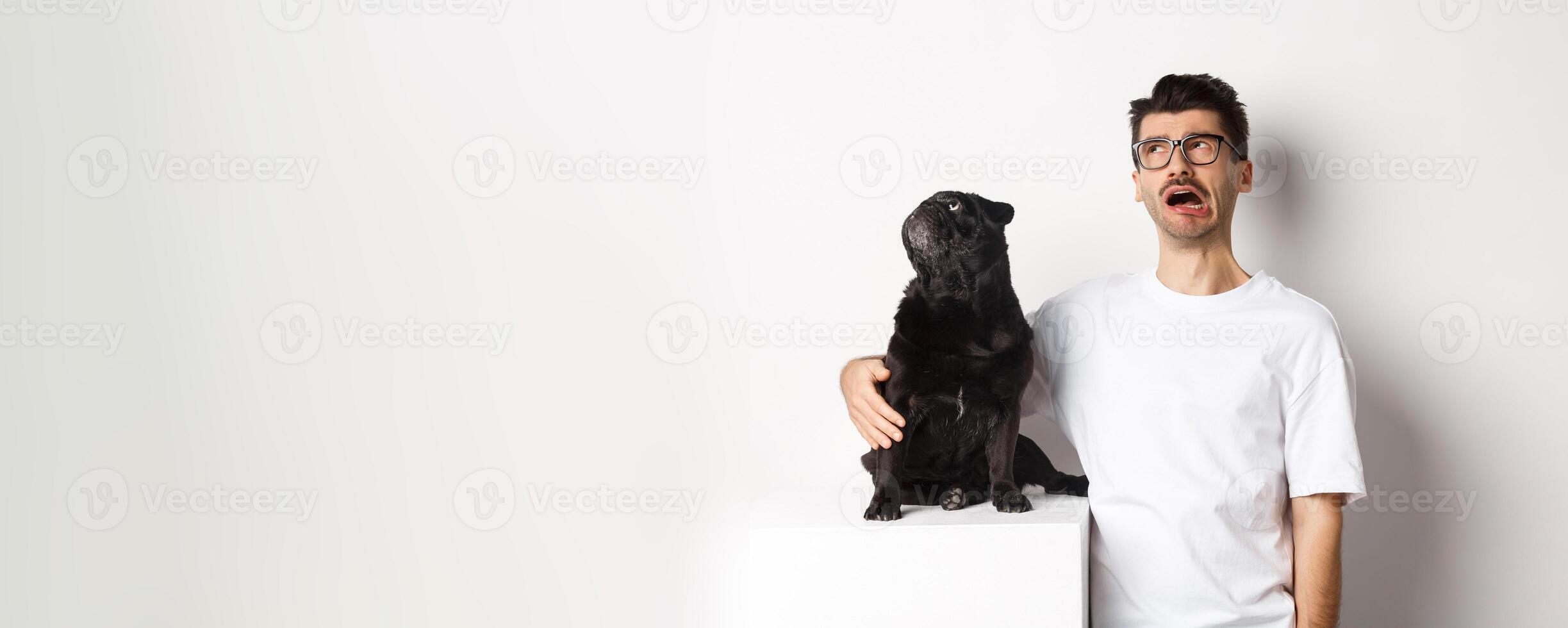 Whining man looking sad while pug staring intrigued at upper left corner, standing against white background photo