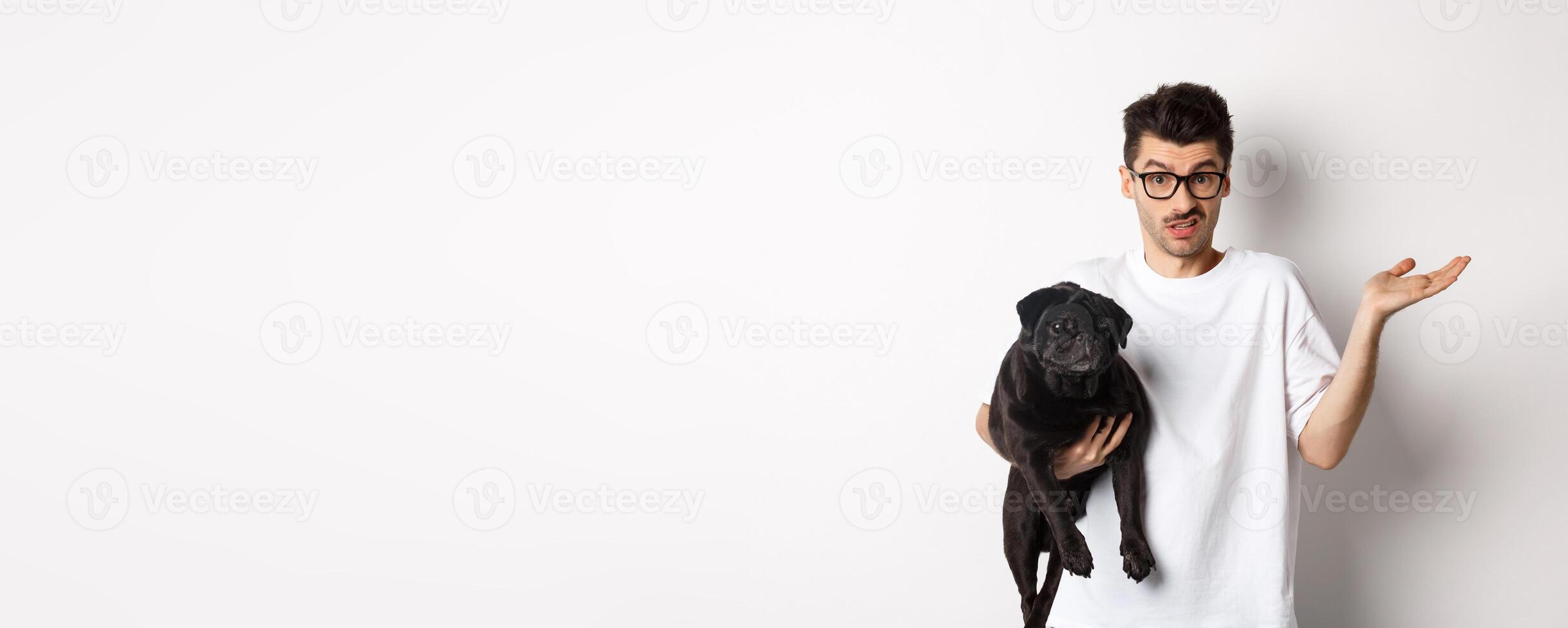 Image of handsome young man holding black pug and looking confused. Guy shrugging shoulders and staring indecisive at camera, carry dog in arm, white background photo