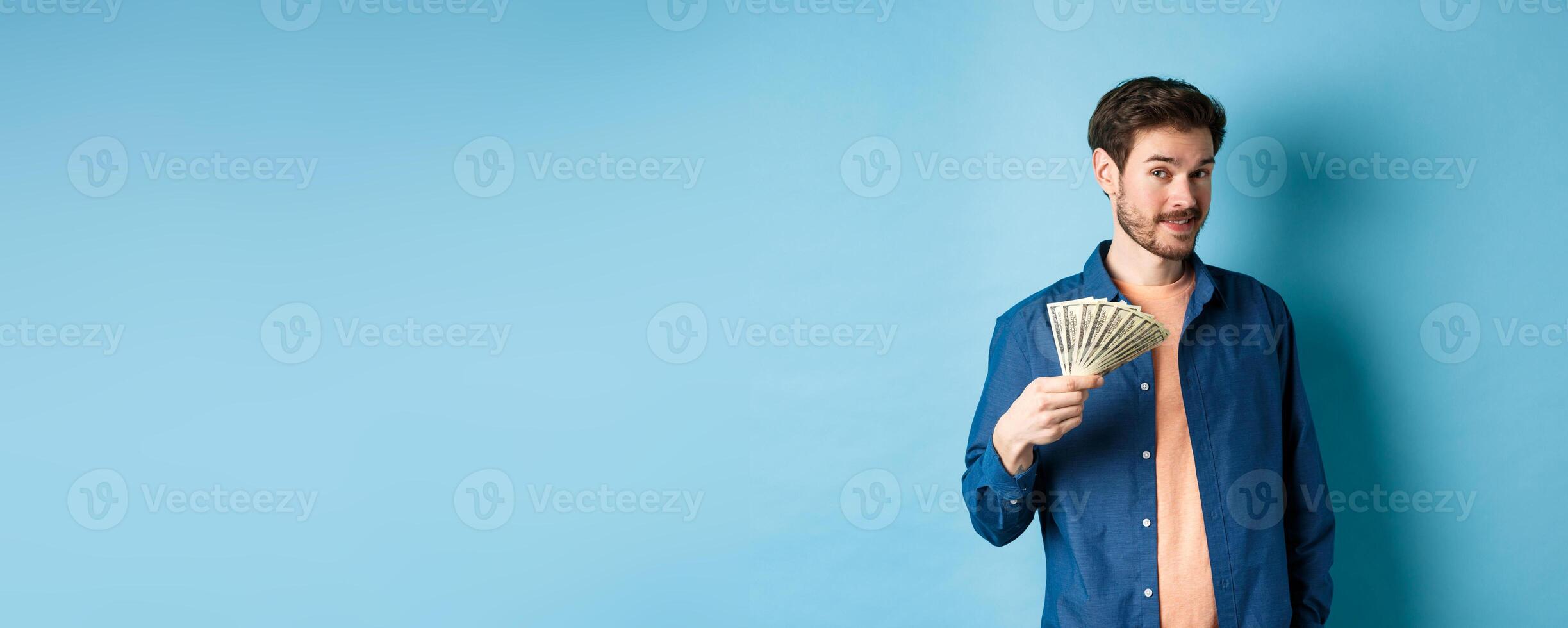 Unbothered rich guy showing cash and smiling, holding dollars in hand, standing on blue background photo