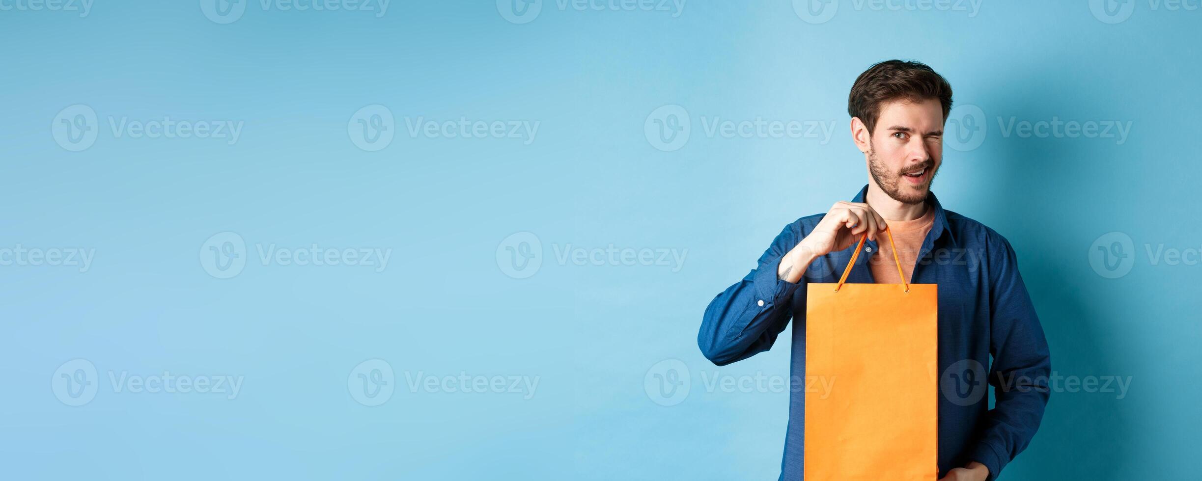 Happy young man showing orange shopping bag and winking, recommending shop discounts, standing on blue background photo