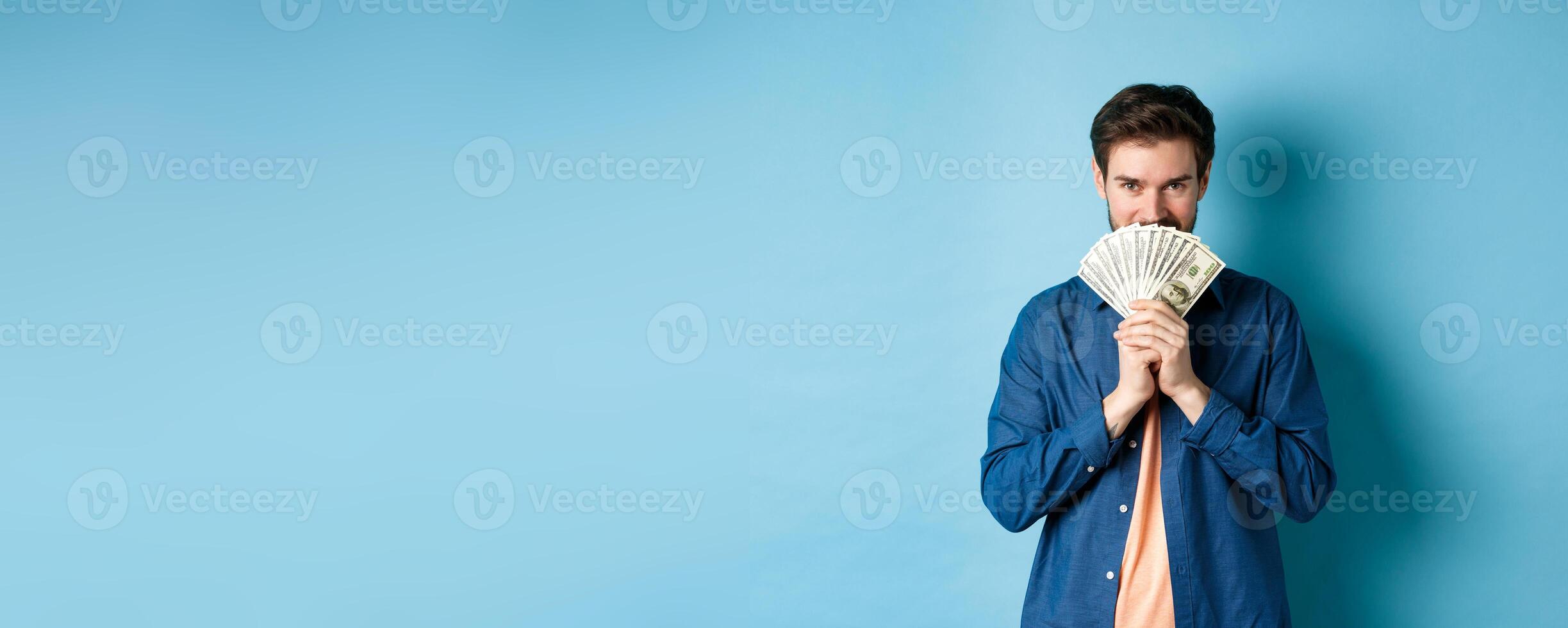 Young man hiding smiling face behind money and looking determined at camera, standing on blue background photo