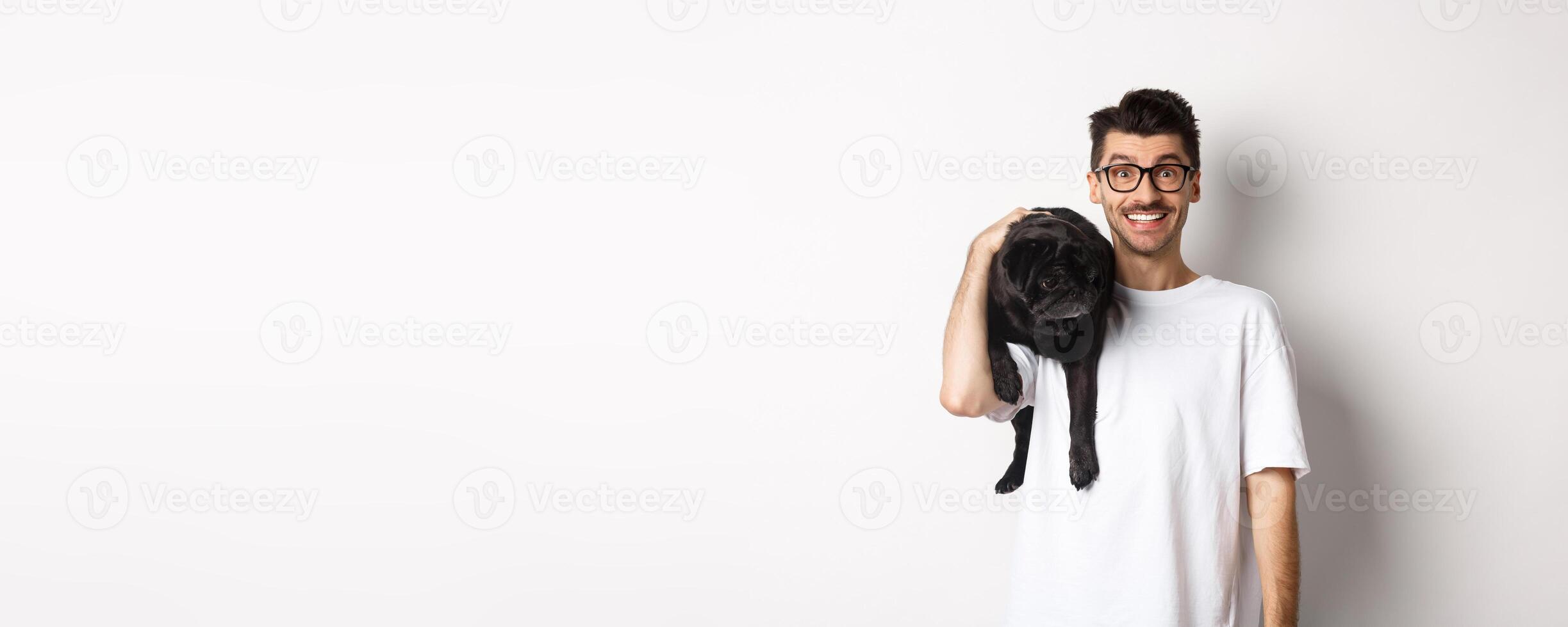 Cheerful hipster man in glasses, holding cute black pug dog on shoulder, staring at camera with happy smile, standing over white background photo