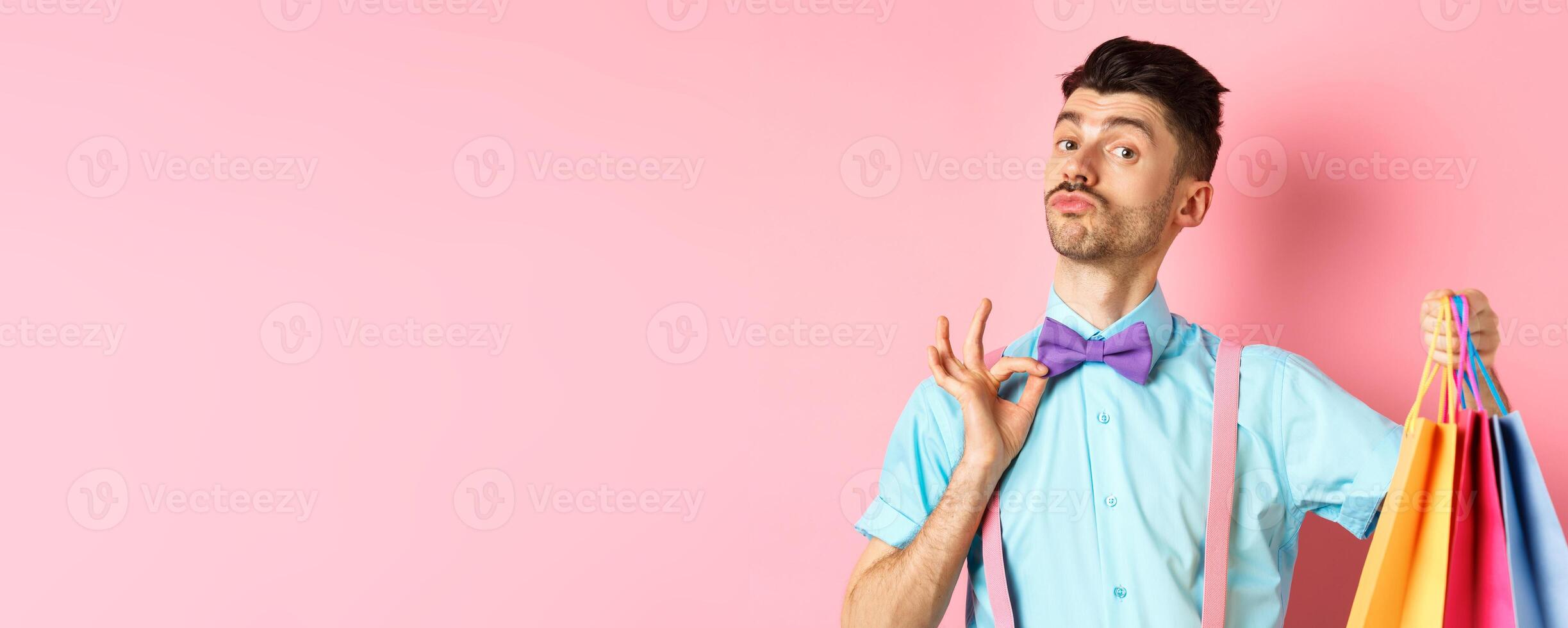 Fancy guy with moustache fixing his bow-tie and holding shopping bags, boyfriend bring gift packages, standing over pink background photo