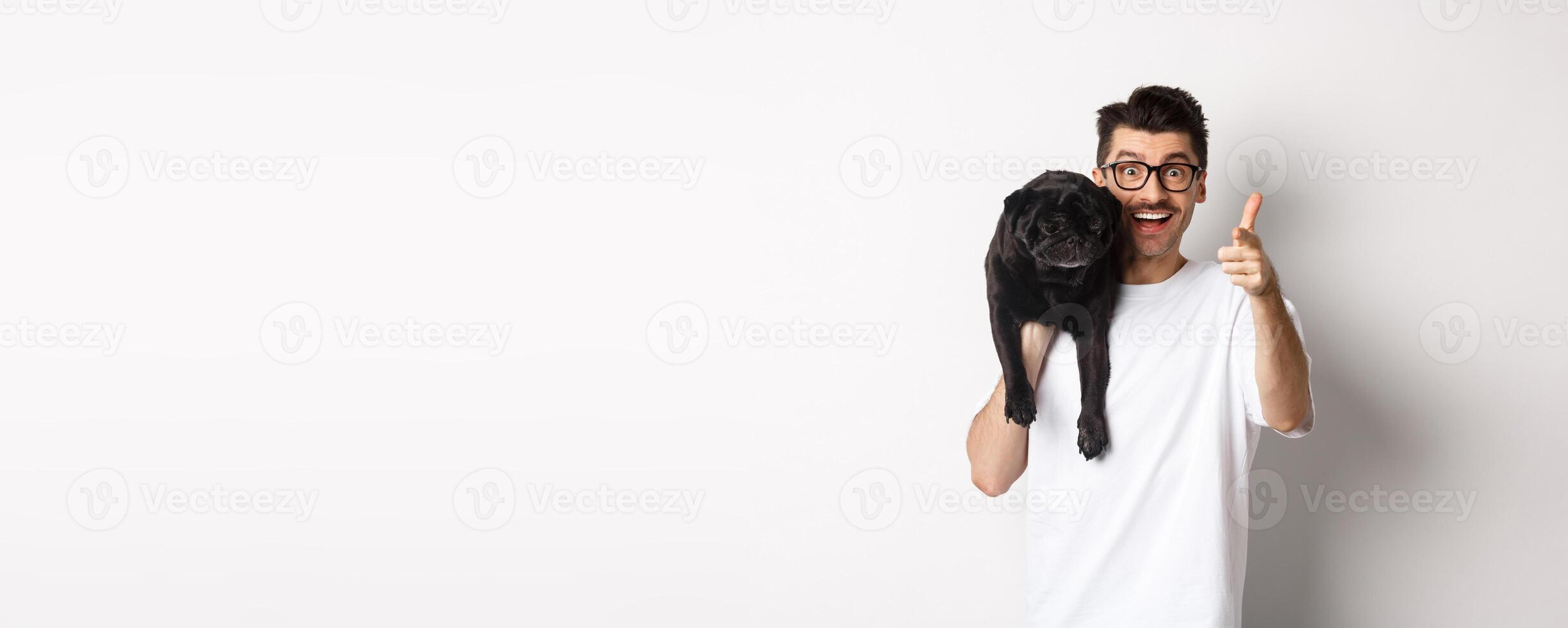 Happy young man holding cute black dog on shoulder and pointing at camera. Hipster guy carry pug on shoulder and staring at camera excited, standing over white background photo