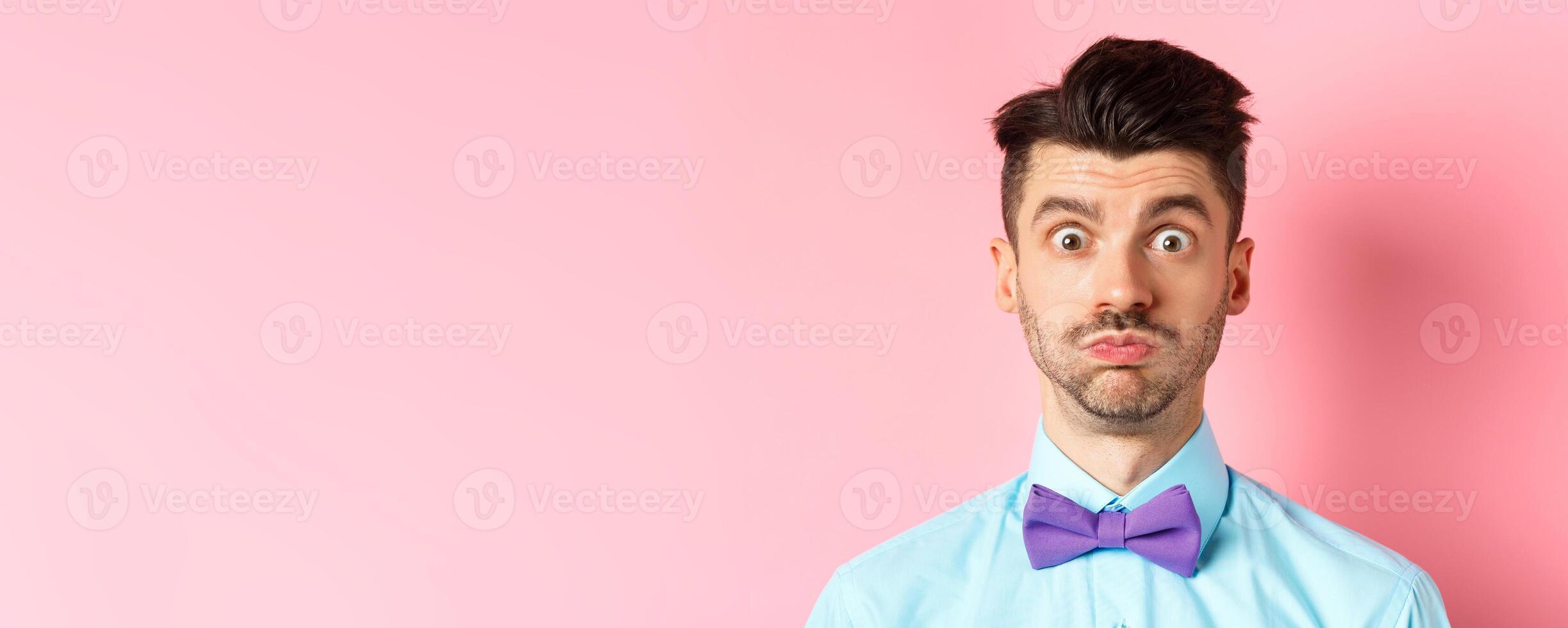 Close-up of confused caucasian guy holding breath, pouting and staring shocked at camera, wearing festive clothes and bow-tie, pink background photo