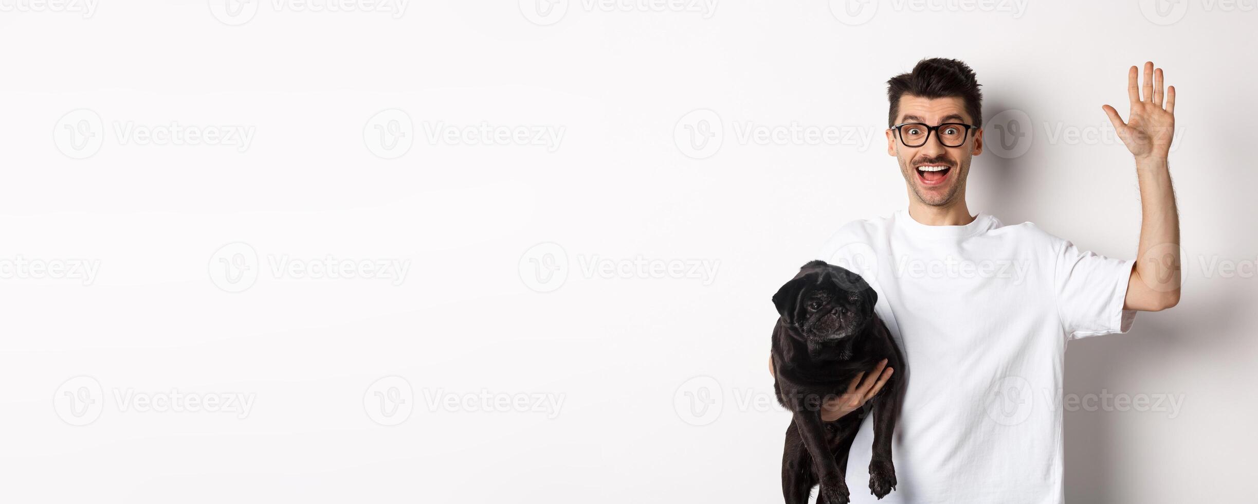 Handsome young man in glasses holding his black pug and waving hand, guy saying hello while carry dog with one arm, standing over white background photo