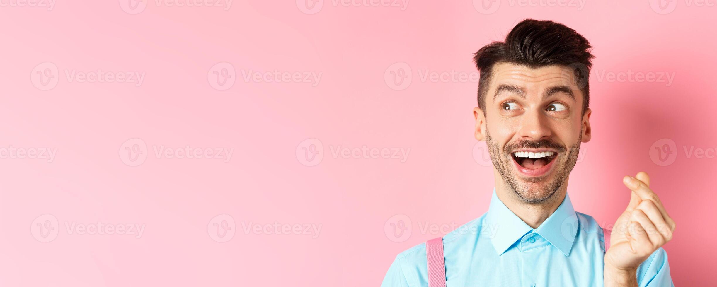 Valentines day concept. Romantic guy showing finger heart and looking left, smiling happy, standing on pink background dreamy photo