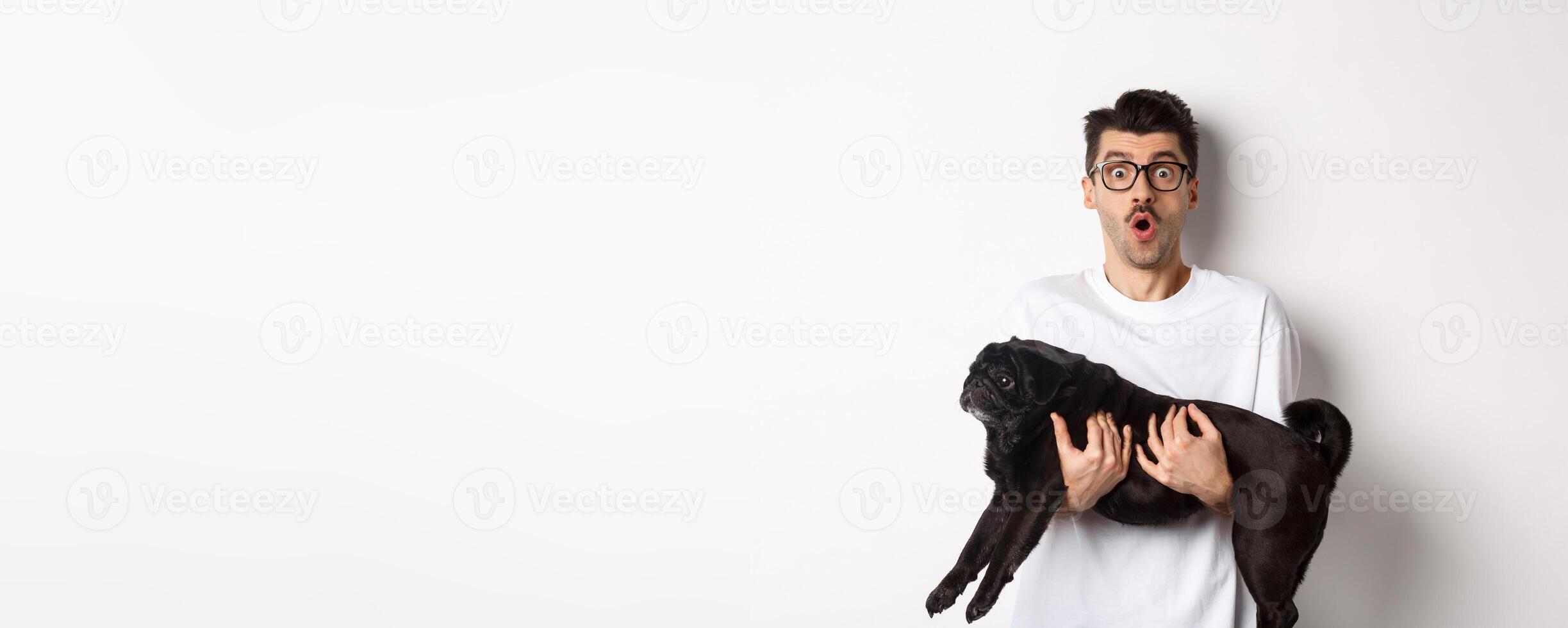 Surprised young man in glasses holding cute black pug, dog owner staring at camera with impressed face, saying wow, standing over white background photo