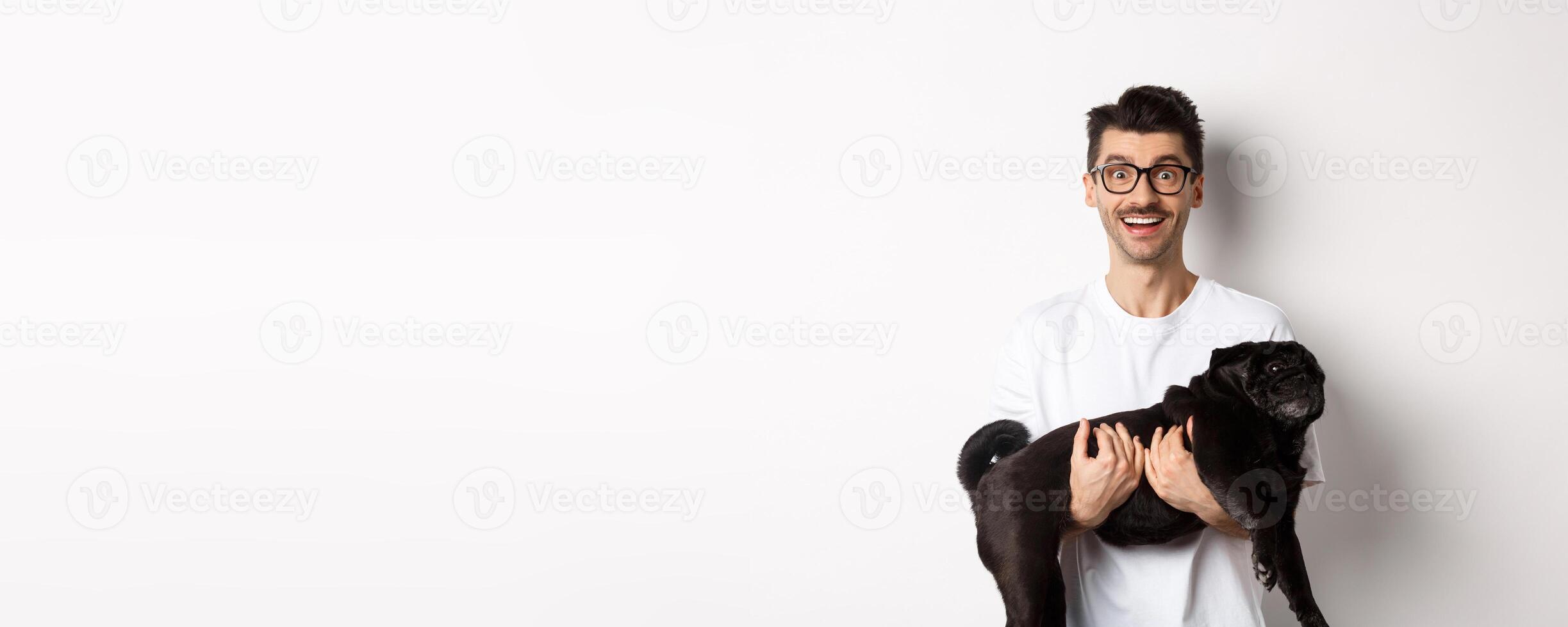 Happy hipster in glasses holding cute black pug and smiling, dog owner staring at camera with amazed smile, standing over white background photo