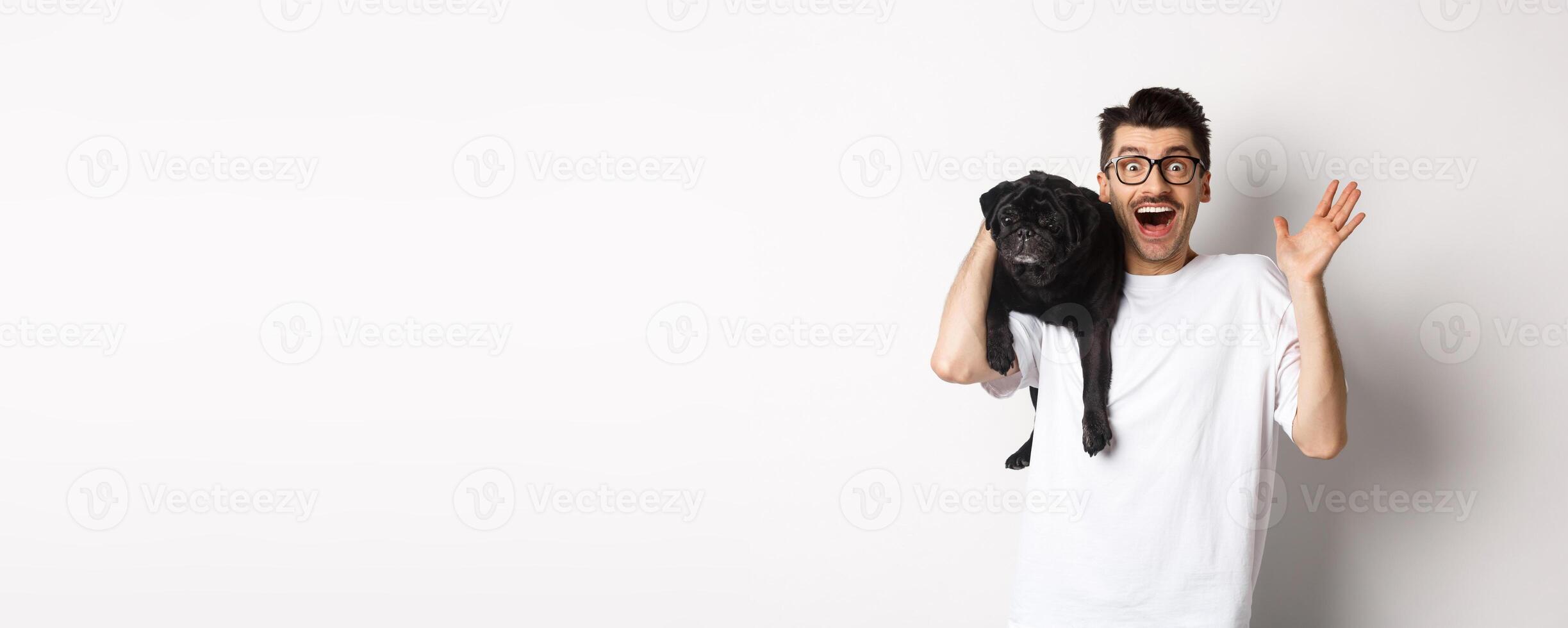 Excited hipster guy in glasses holding dog on shoulder, waving hand to say hello, greeting someone with happy face. Happy man carry his cute pug and smiling, white background photo