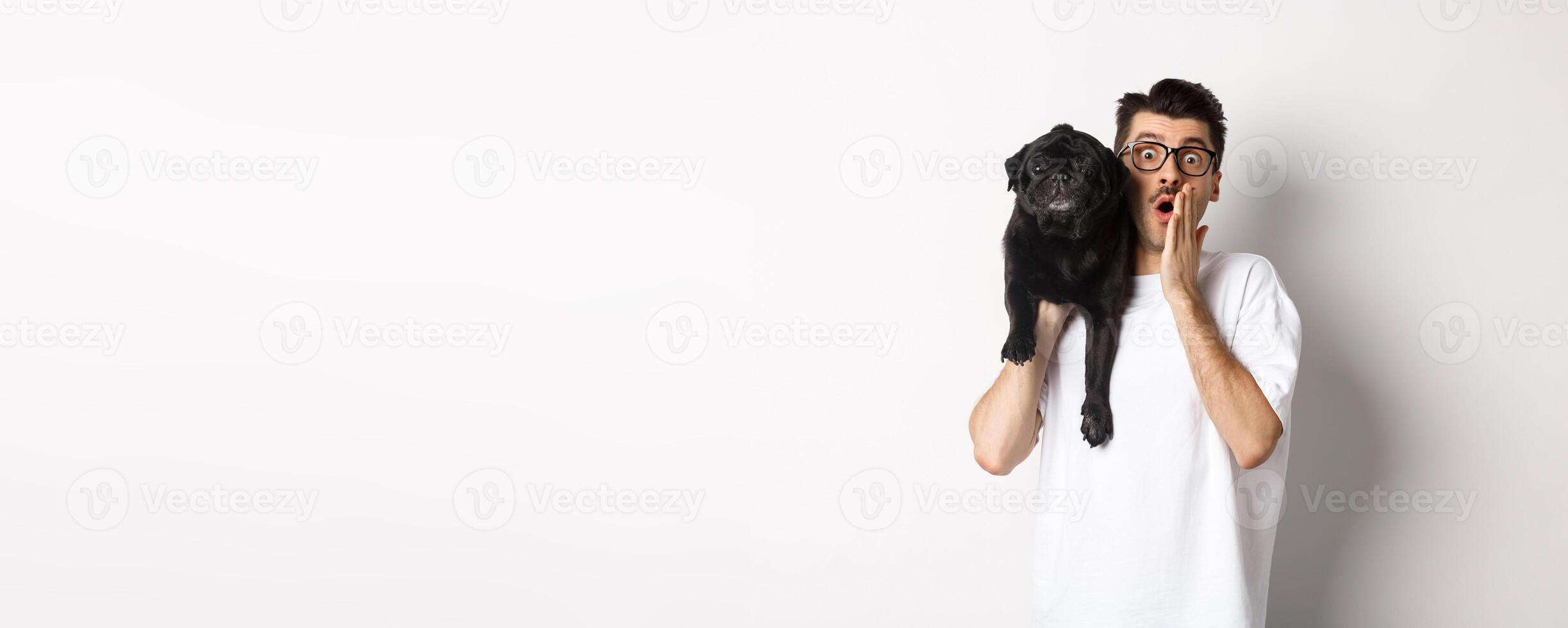 Image of shocked dog owner staring at camera and gasping impressed, cute black pug sitting on his shoulder and staring at you, white background photo