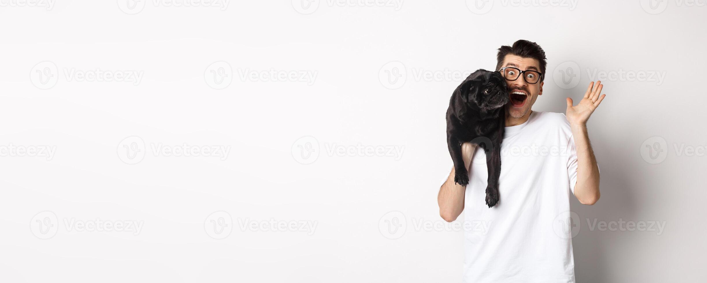 Handsome young man in glasses holding his black pug and waving hand, guy saying hello while carry dog with one arm, standing over white background photo
