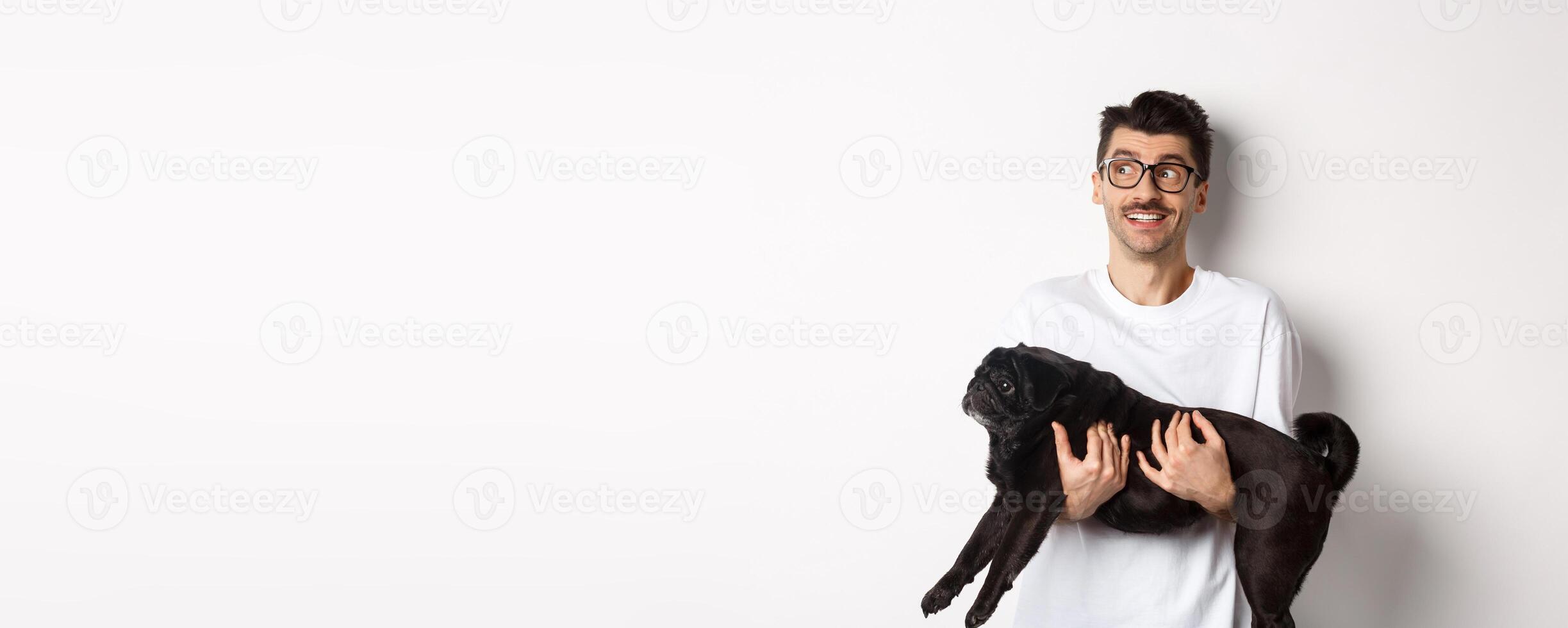 Excited hipster guy with his dog looking left at logo. Handsome pet owner carry black pug and staring at promo banner, standing over white background photo