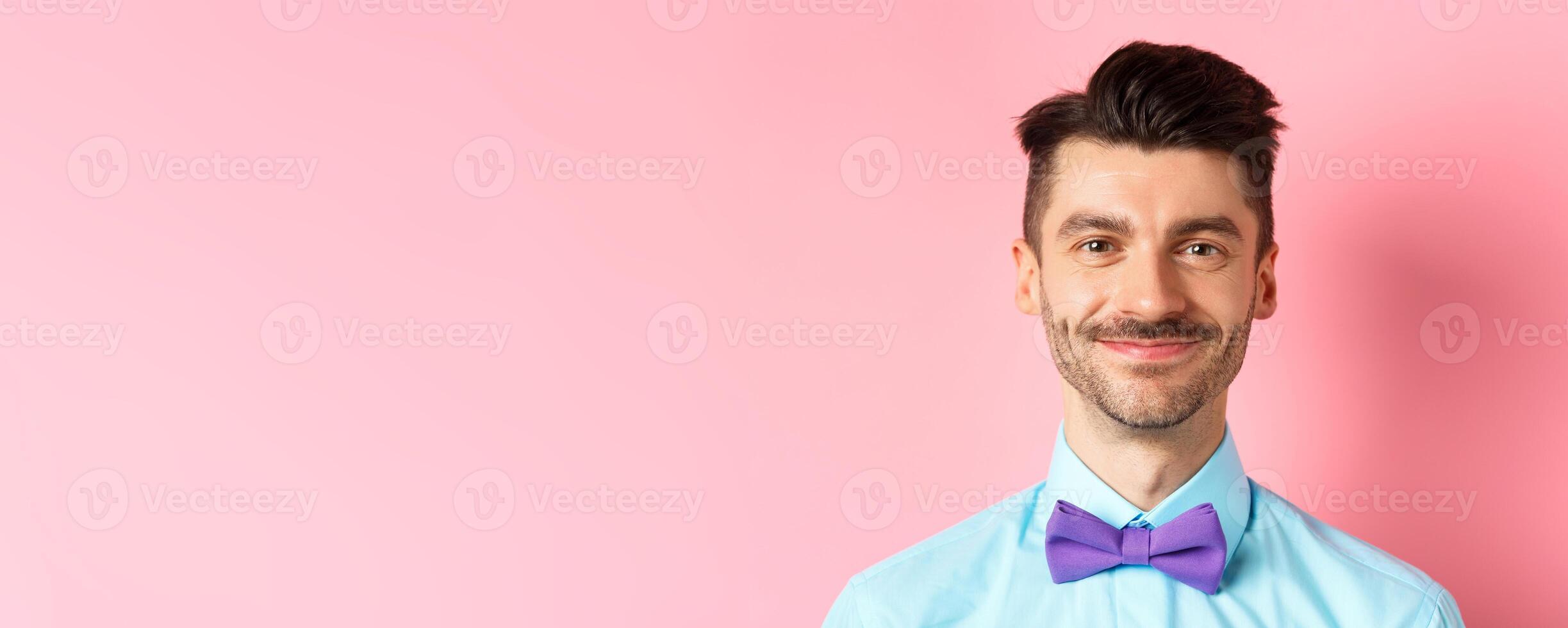 Close-up of smiling hansome man with moustache, wearing bow-tie and shirt, standing cheerful on pink background photo
