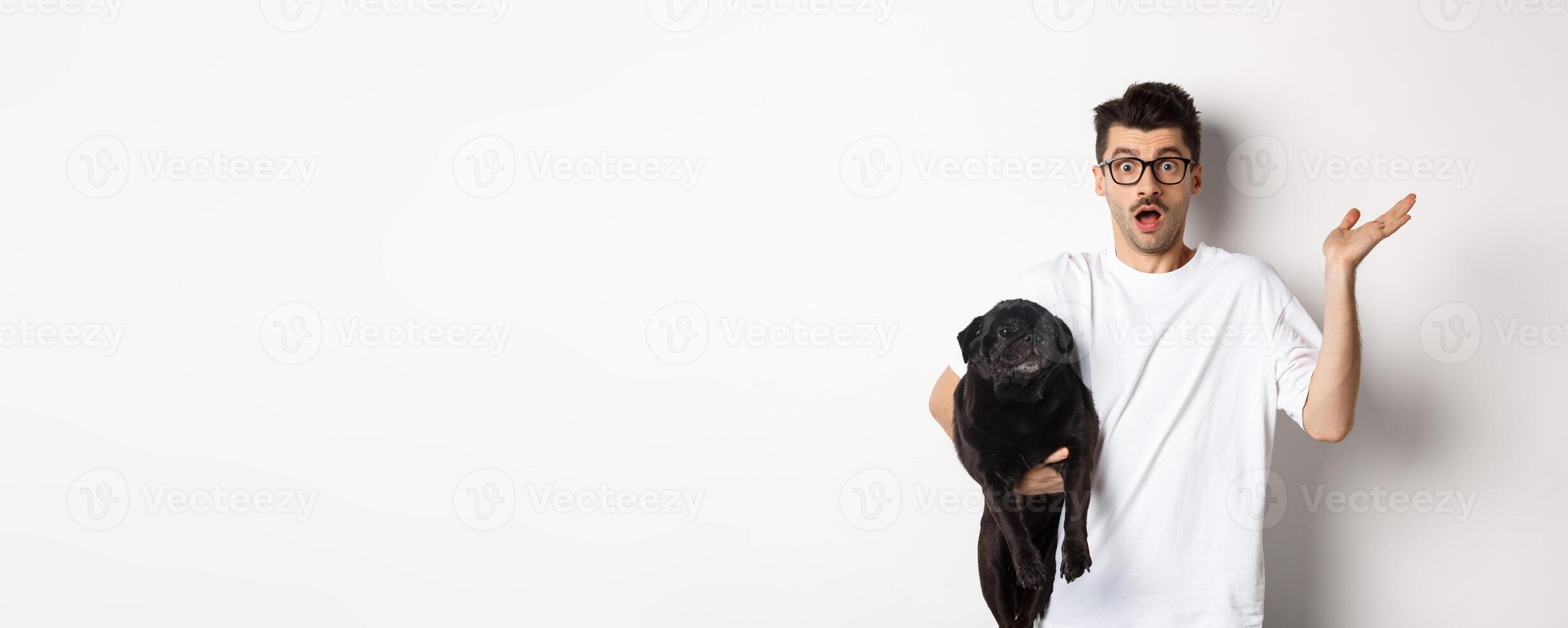 Image of confused hipster man holding dog and shrugging, dont know, raising hand puzzled, standing with his animal over white background photo