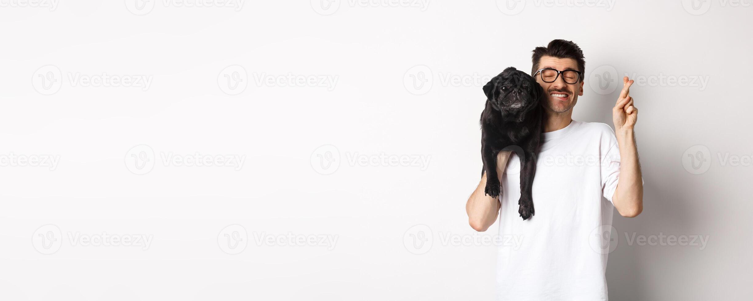 Hopeful smiling dog owner making a wish, holding cute black pug on shoulder and cross fingers for good luck, white background photo