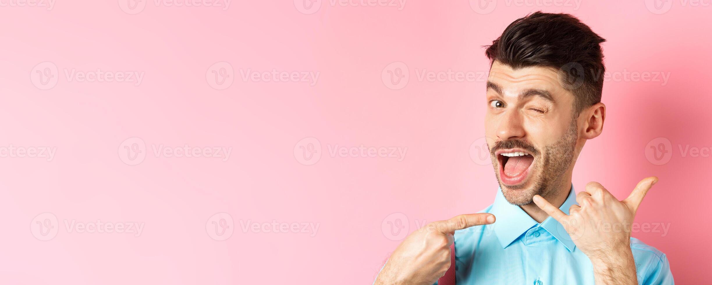 Cheeky and funny guy showing phone gesture, asking to call him and winking, telling dial his number, standing over pink background photo