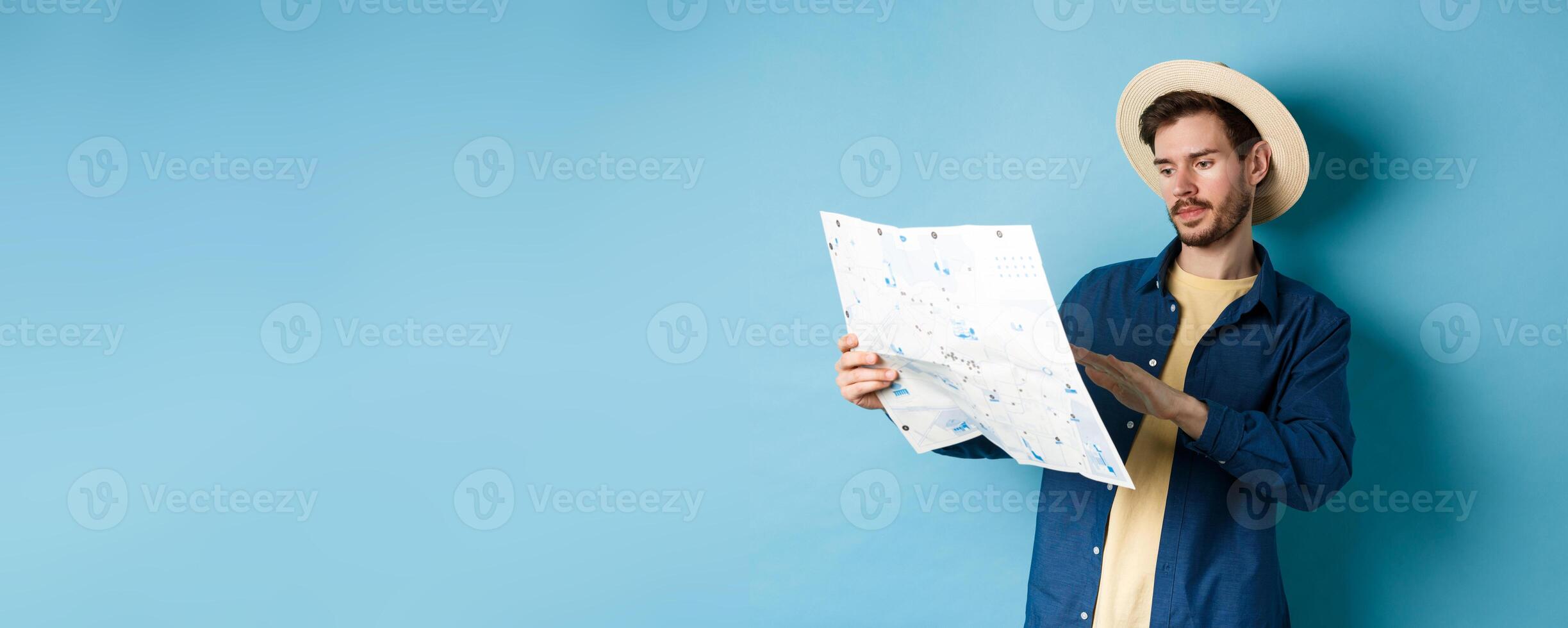 Image of handsome caucasian man looking at travel map, studying route during summer vacation, standing in straw hat on blue background photo