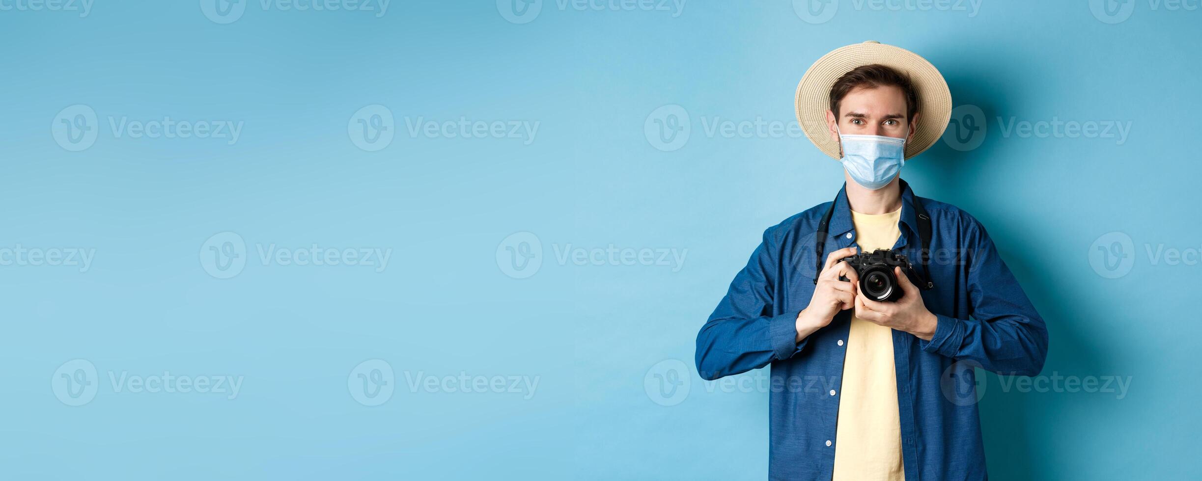 Covid-19, pandemic and travel concept. Happy guy on vacation taking pictures with camera, wearing medical mask and summer hat, blue background photo