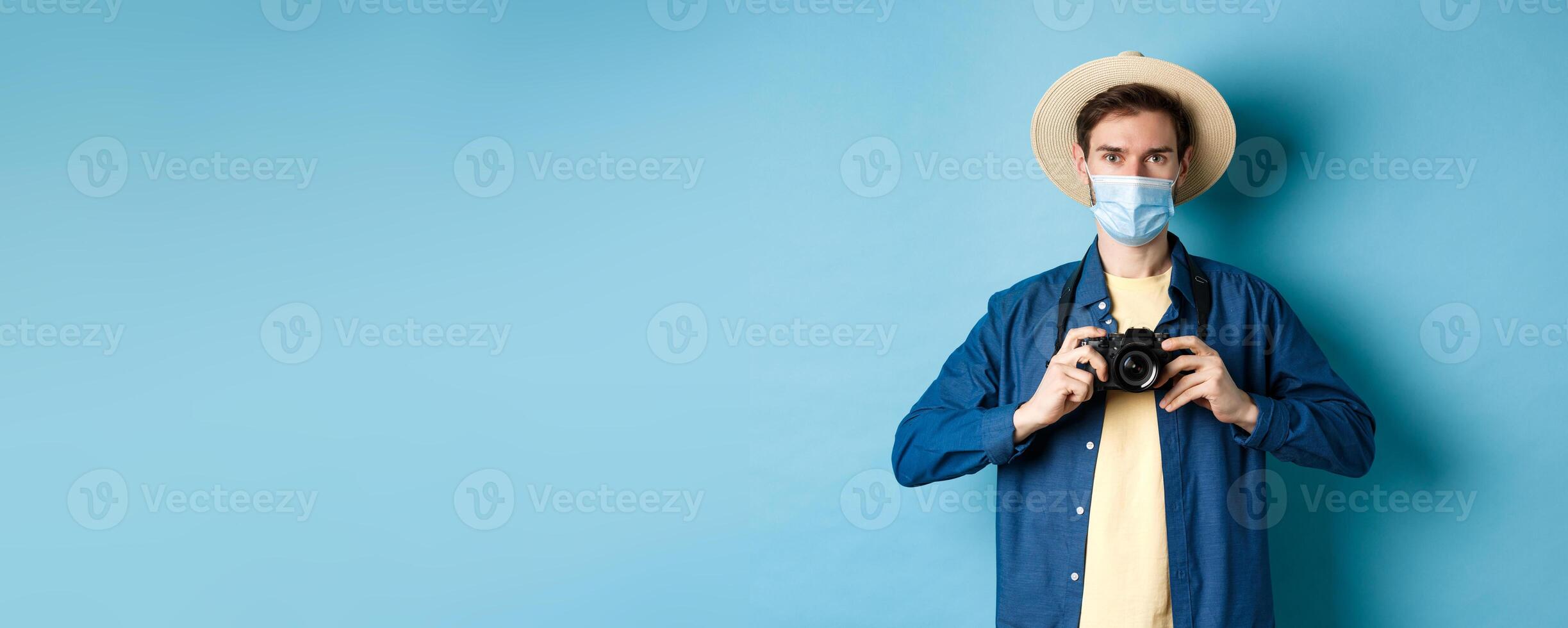 Covid-19, pandemic and travel concept. Cheerful tourist in summer hat and medical mask taking photos, holding camera, standing on blue background photo