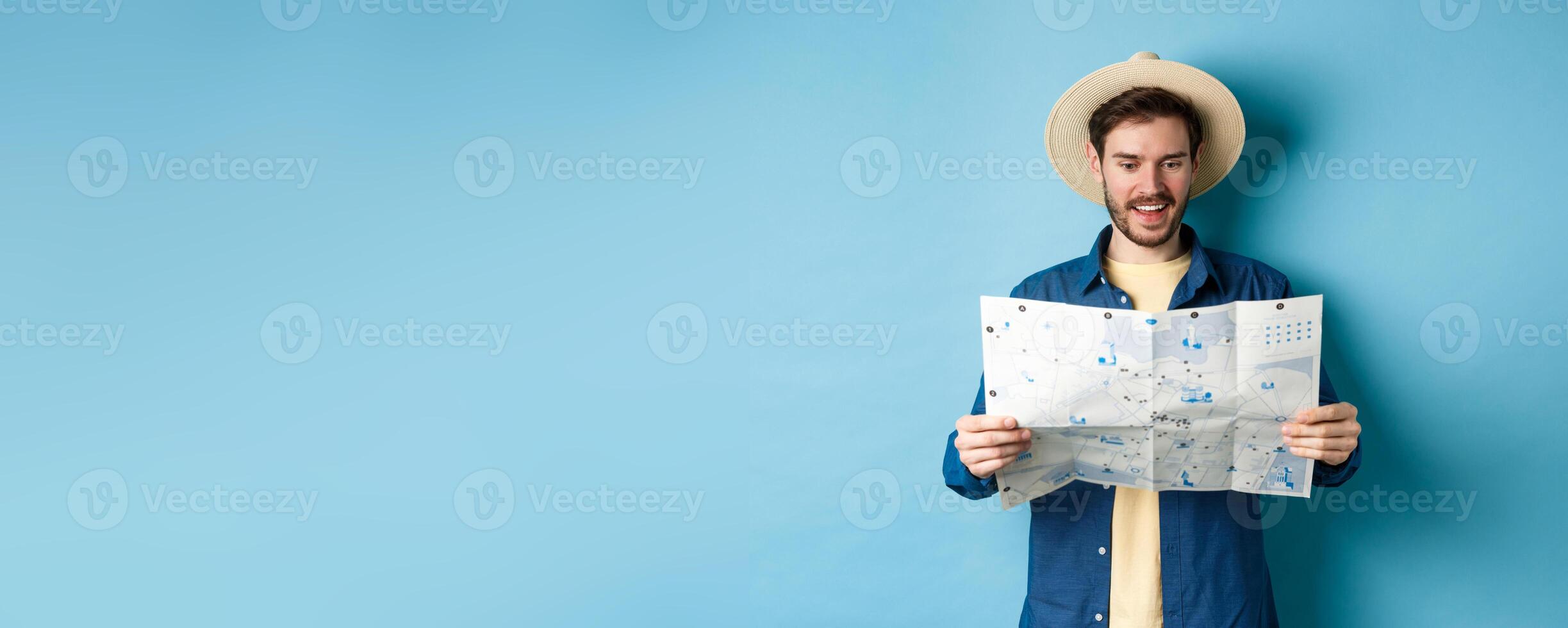 Cheerful tourist in summer hat planning travel route on vacation, looking at sighseeing map and smiling excited, standing on blue background photo