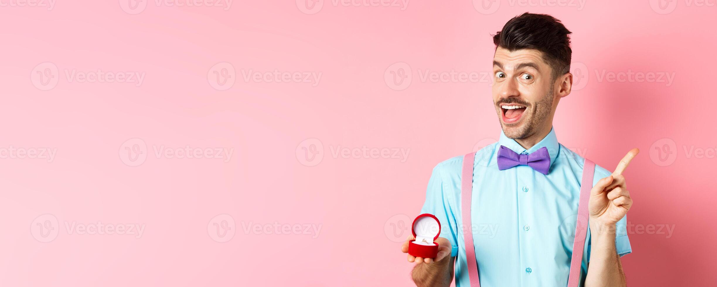 Valentines day. Funny guy with moustache and bow-tie, showing engagement ring and pointing finger at upper right corner, standing on pink background photo