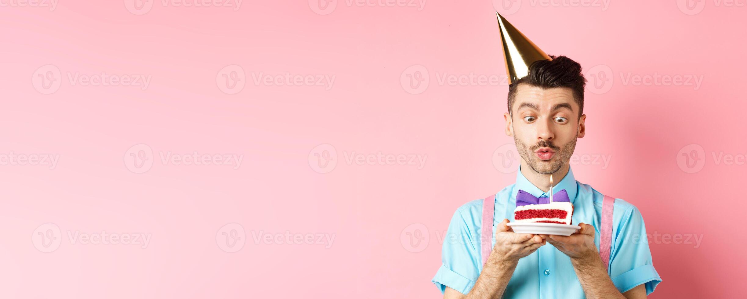 Holidays and celebration concept. Happy young man in birthday party hat blowing candle on cake, making wish on b-day, standing over pink background photo