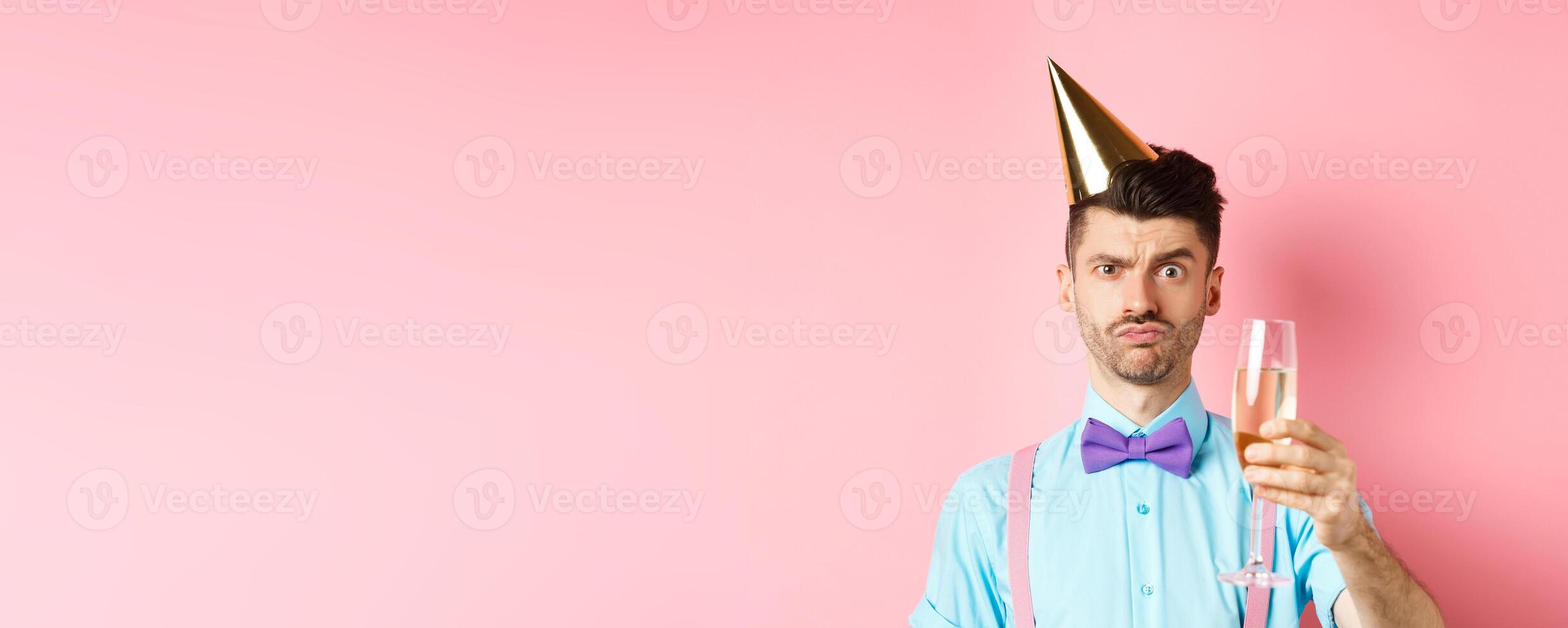 Holidays and celebration concept. Troubled young man in party hat, frowning with doubtful face, raising glass of champagne perplexed, standing on pink background photo