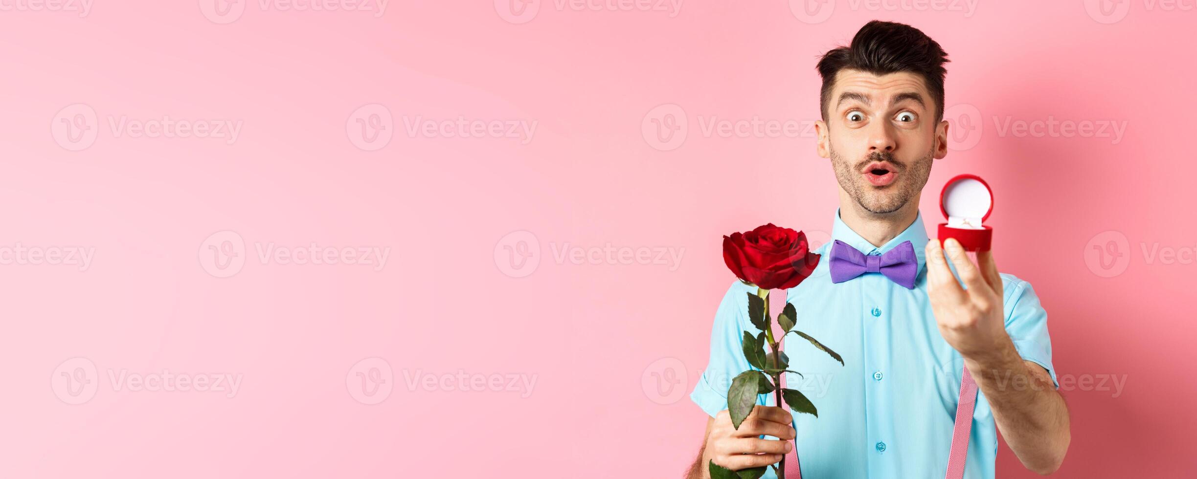 Valentines day. Funny man with moustache and bow-tie making proposal, showing engagement ring and propose with red rose, standing over pink background photo