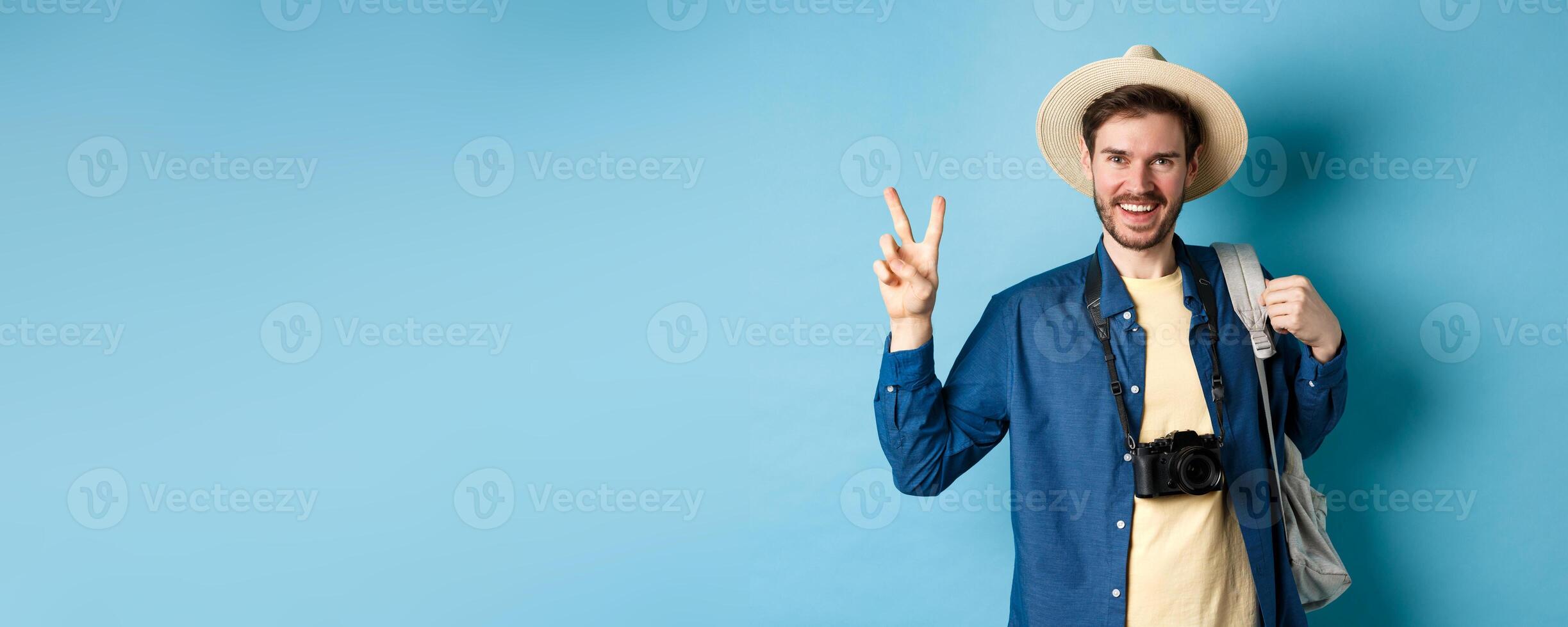 Happy handsome man taking photo on summer vacation, showing peace sign and smiling, wearing straw hat and holding tourist backpack, blue background