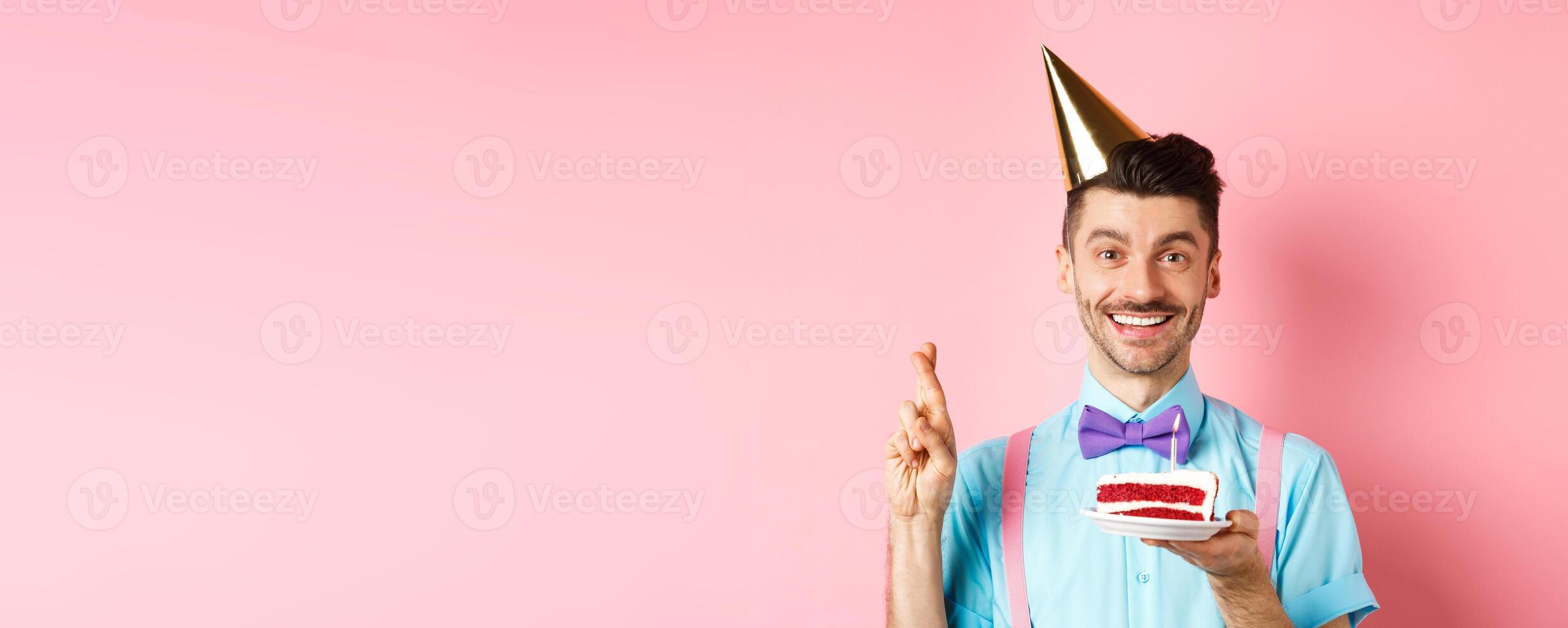 Holidays and celebration concept. Happy young man enjoying birthday party, wearing cone hat and cross fingers, making wish on bday cake with candle, pink background photo