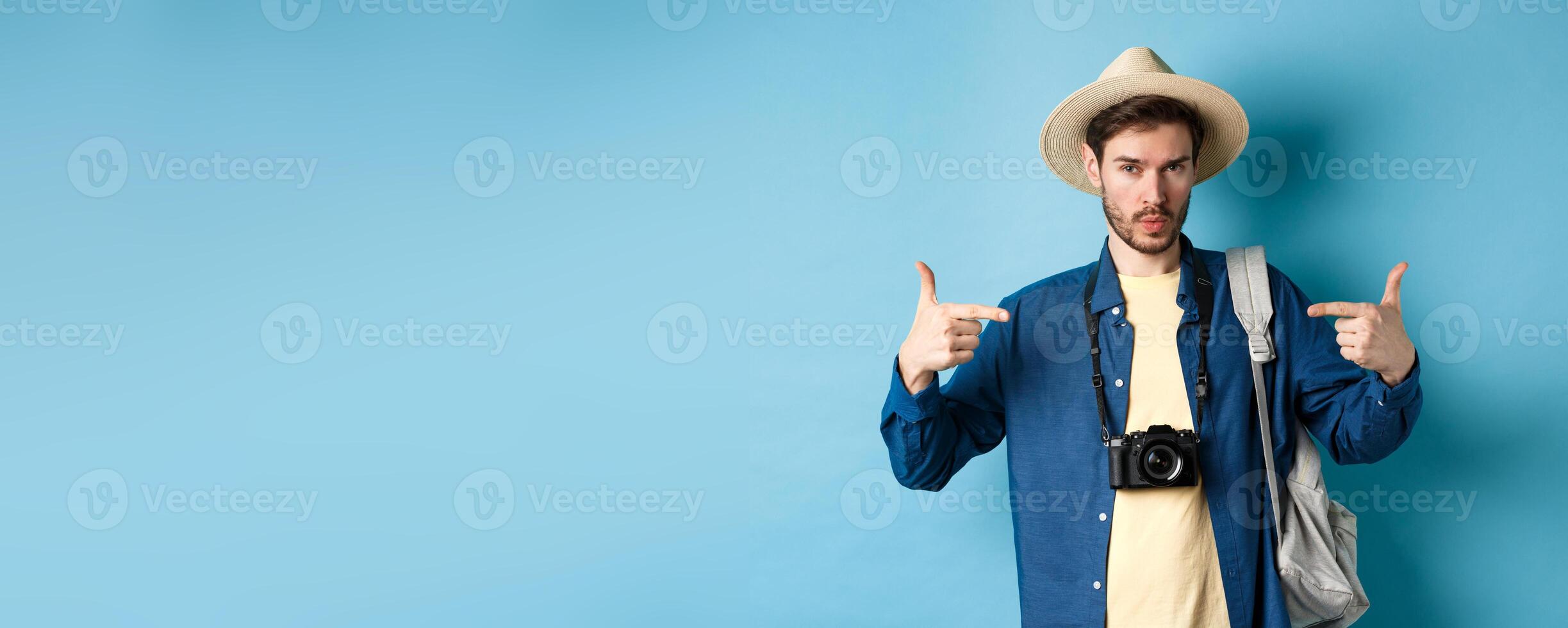 Confident and ready young man pointing at himself, self-promoting or show-off, going on summer vacation, wearing straw hat, holding backpack with camera, blue background photo