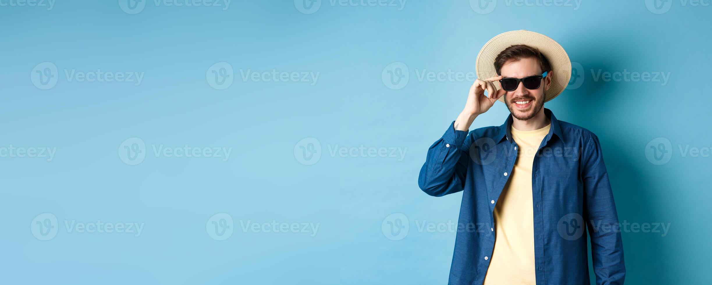 Happy smiling guy going on summer vacation, wearing straw hat and black sunglasses, standing on blue background photo