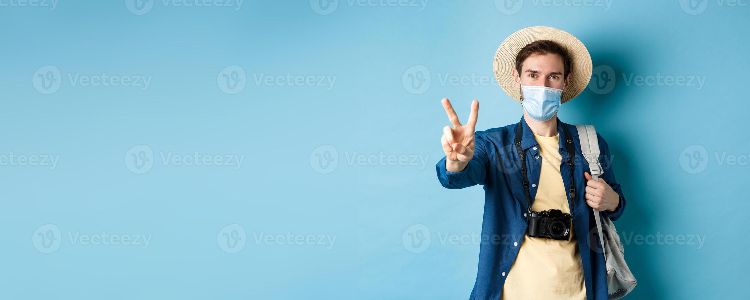 Covid-19, pandemic and travel concept. Positive guy going on vacation, wearing summer hat and medical mask, showing peace sign, standing with backpack and camera on blue background photo