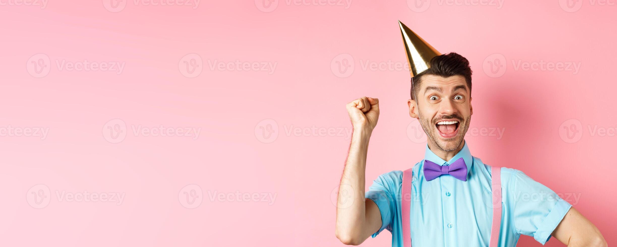 Holidays and celebration concept. Happy birthday boy with moustache and bow-tie celebrating event in party cone hat and festive clothes, raising hand up and shout yes photo