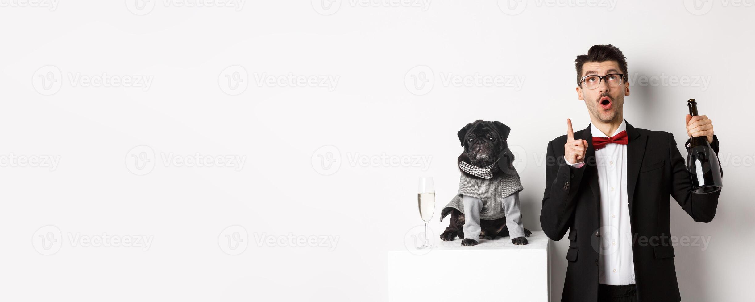 Pets, winter holidays and New Year concept. Handsome young man in suit celebrating Christmas with black dog, puppy wearing costume, owner looking and pointing up at copy space photo
