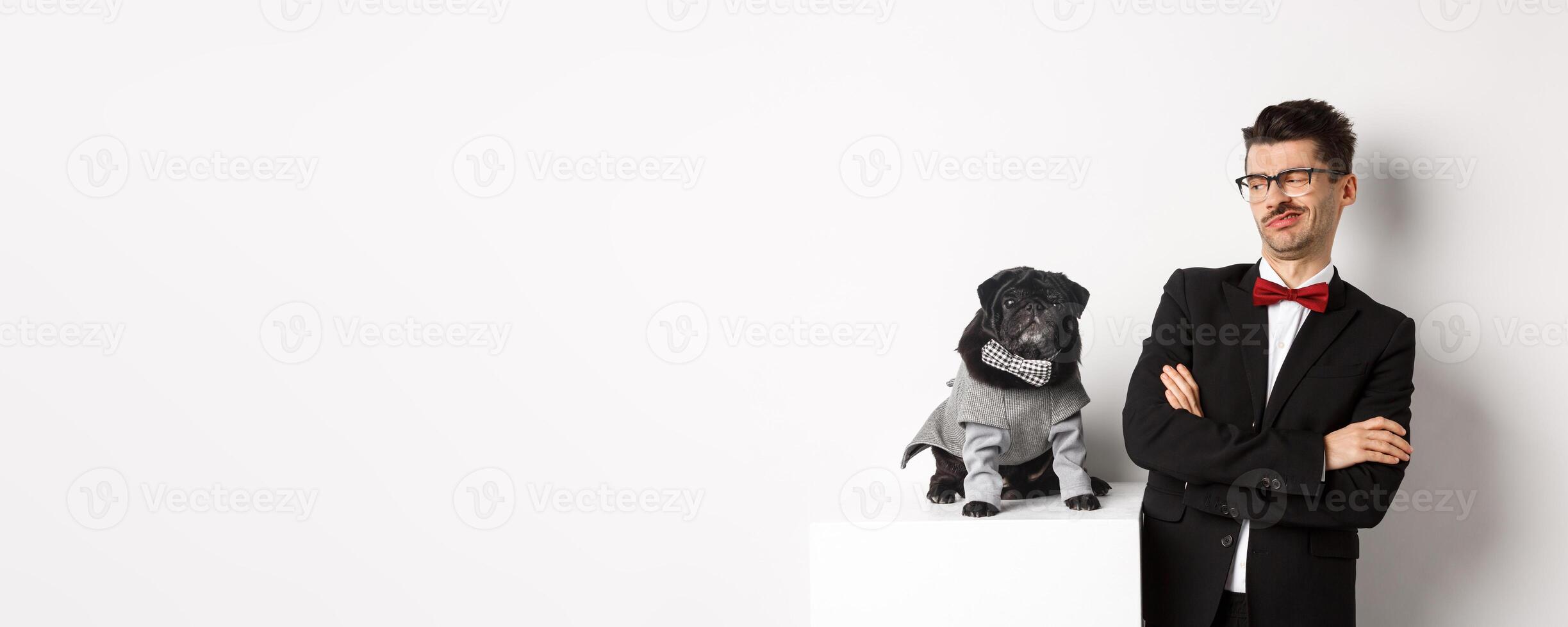 Animals, party and celebration concept. Image of funny young man in suit and glasses, looking skeptical at cute pug in costume, standing over white background photo