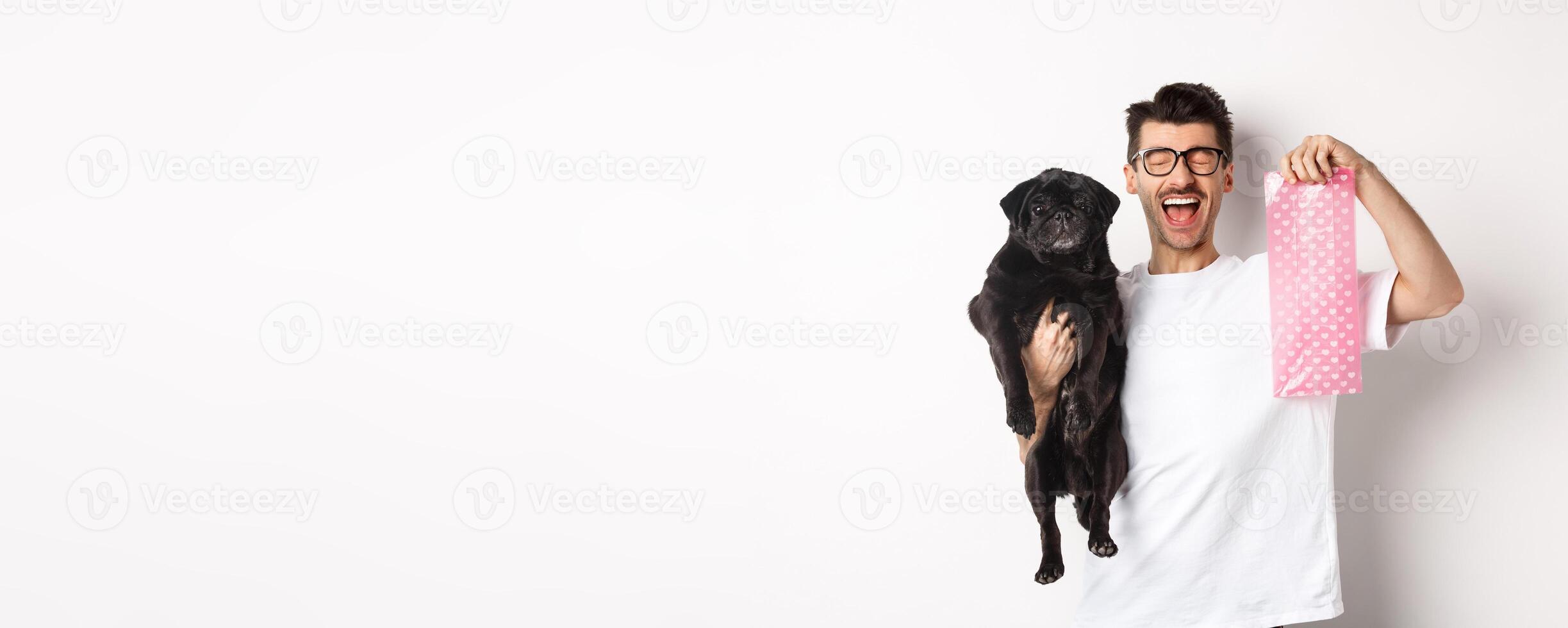 Image of happy man, pet owner, holding cute black pug and dog poop bag, standing over white background photo