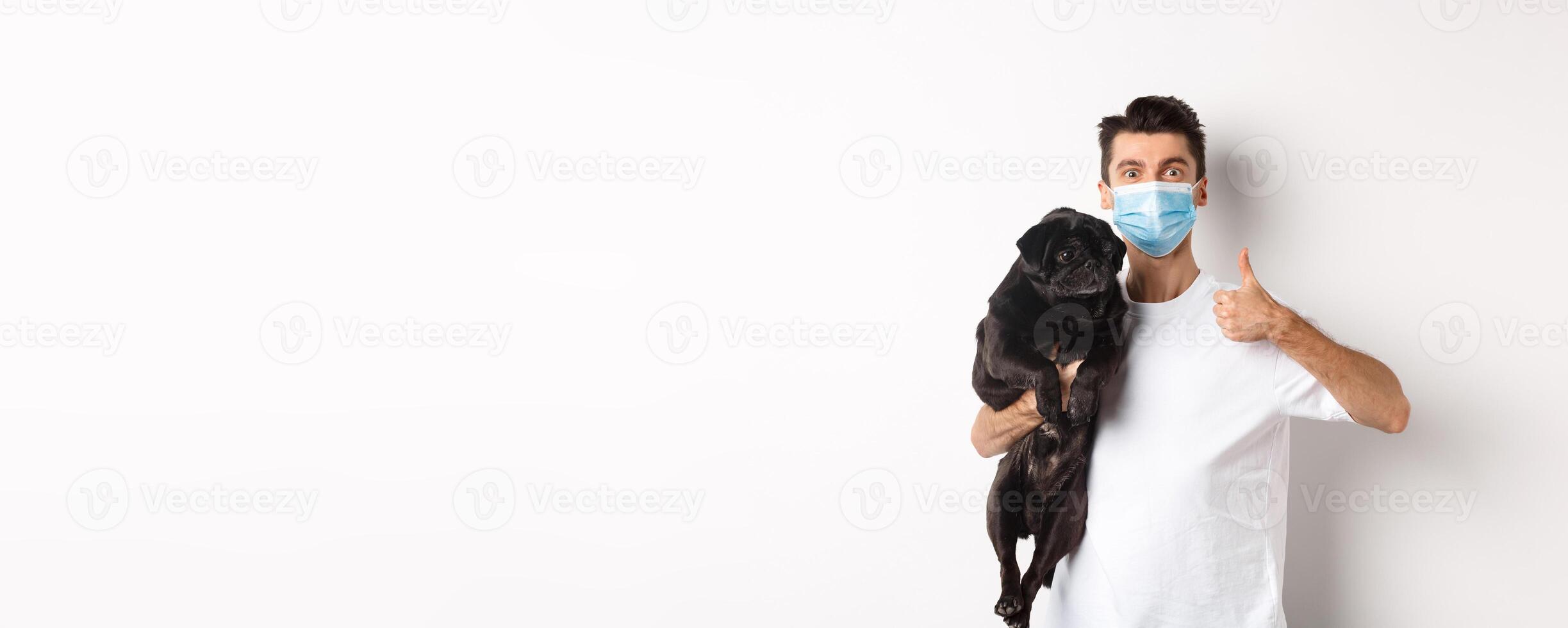 Covid-19, animals and quarantine concept. Young man in medical mask holding cute black pug dog, showing thumb up, like and approve, standing over white background photo