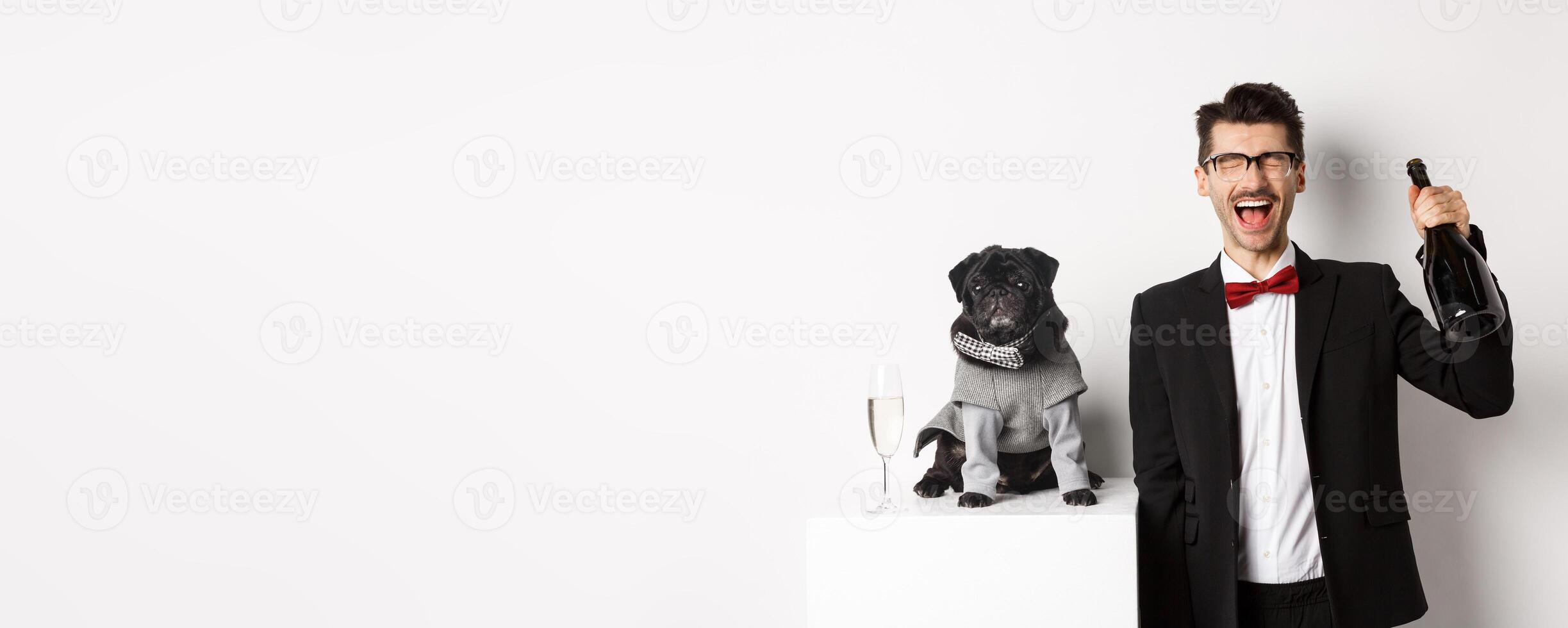Pets, winter holidays and New Year concept. Happy man celebrating Christmas party pet, standing with cute dog in costume, drinking champagne and rejoicing, white background photo
