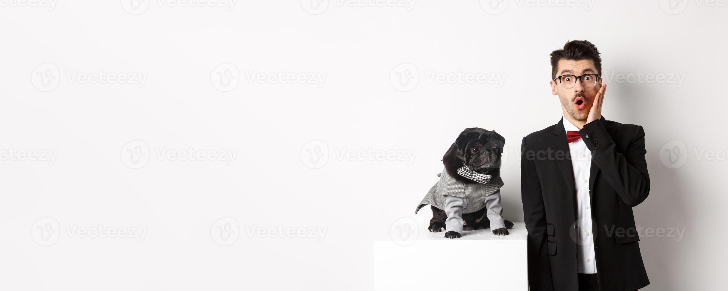 Animals, party and celebration concept. Shocked handsome man in formal suit and cute dog in costume, staring at camera amazed, standing over white background photo