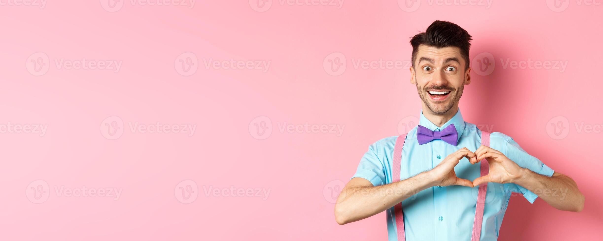 Passionate guy in funny bow tie saying I love you, showing heart gesture on Valentines day and smiling, expressing sympathy to lover, standing over pink background photo
