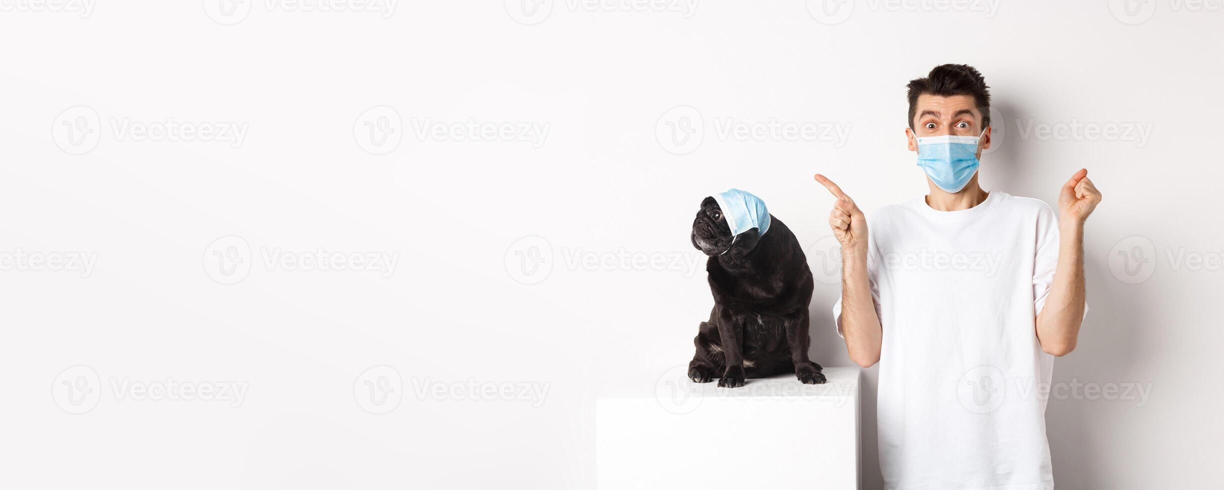 Covid-19, animals and quarantine concept. Young man and black dog wearing medical masks, pug looking at upper left corner and owner showing promo offer, rejoicing photo