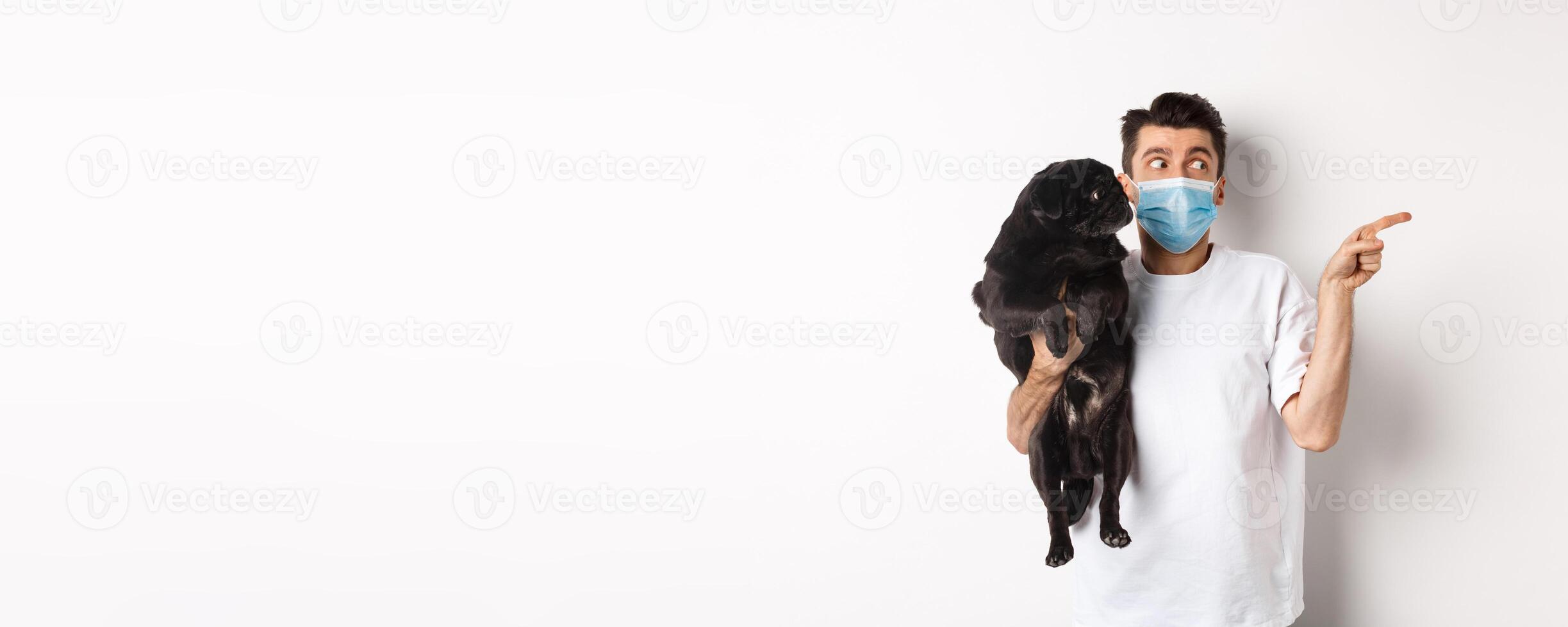 Covid-19, animals and quarantine concept. Young man in face mask holding cute black pug, dog and pet owner looking right at copy space, standing over white background photo
