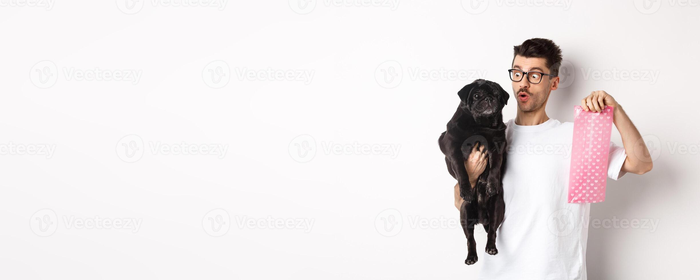 Cheerful young man holding black pug and pink dog poop bag, standing over white background photo