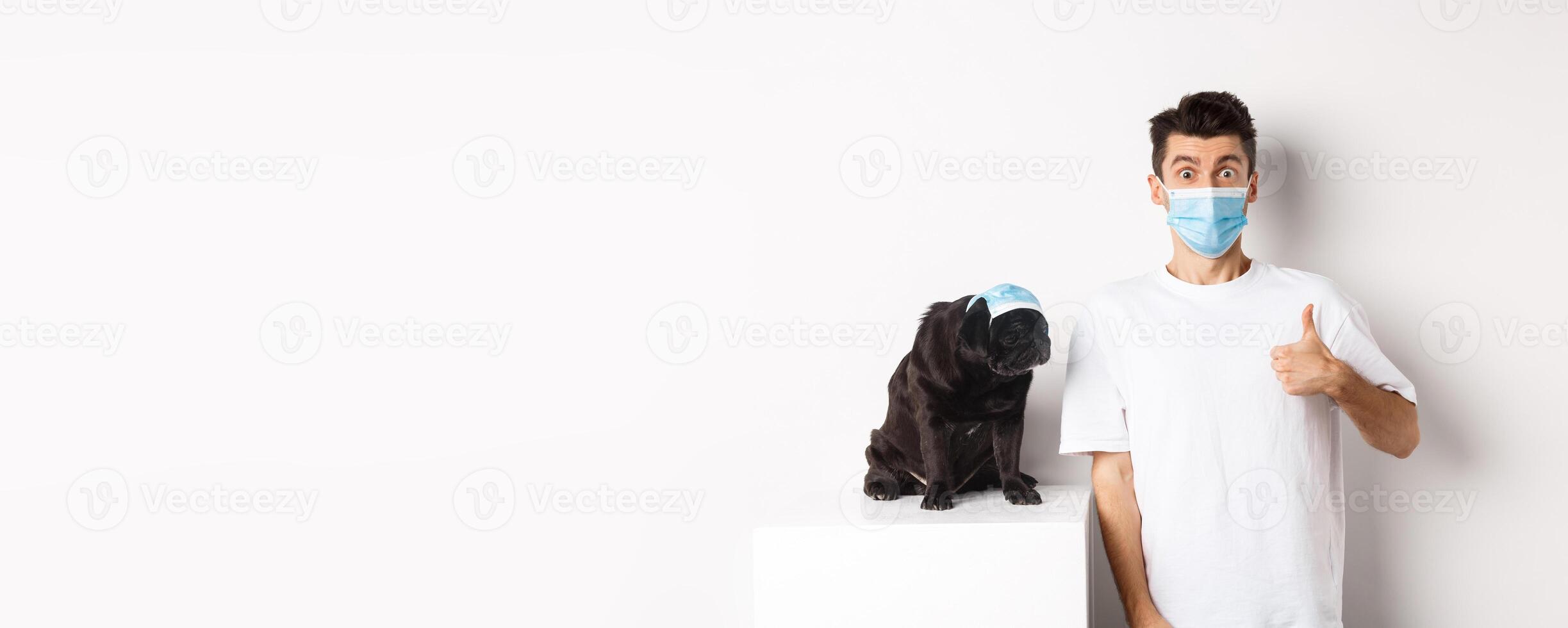 Covid-19, animals and quarantine concept. Handsome young man and small dog wearing face masks, owner showing thumb up in approval, praise super cool product, white background photo