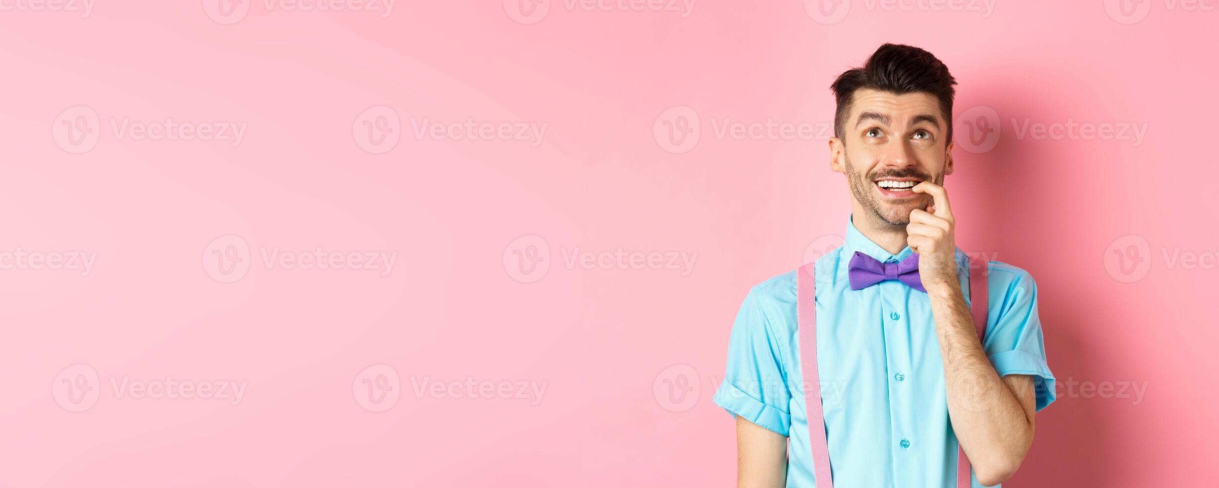 Image of smiling man making choice, looking dreamy and happy up, biting fingernail with tempted expression, wanting something, standing over pink background photo