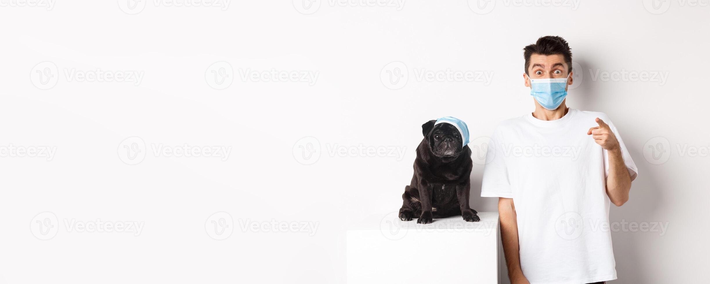 Covid-19, animals and quarantine concept. Happy dog owner and cute pug wearing medical masks, man pointing finger at camera amazed, white background photo