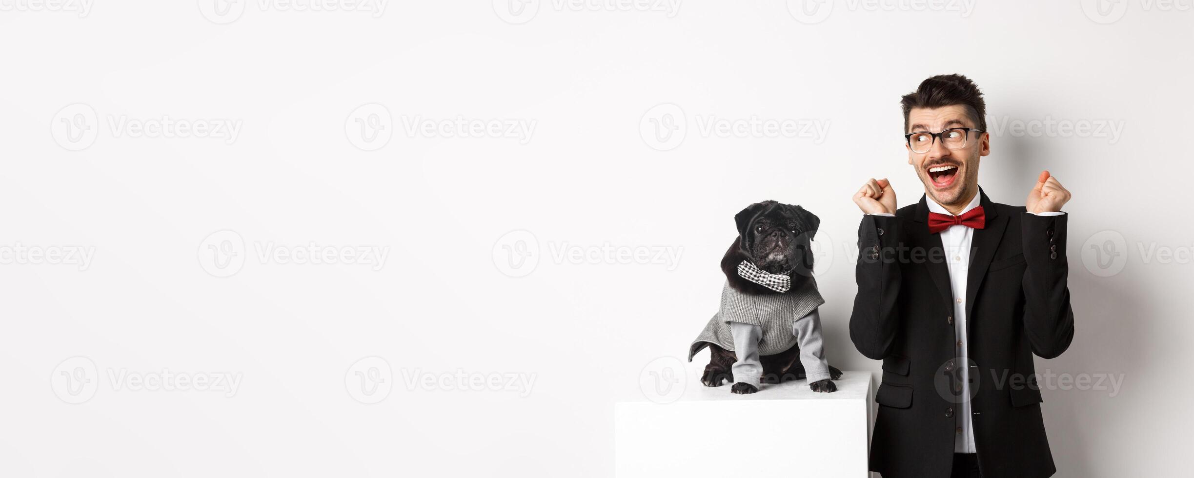 Animals, party and celebration concept. Cheerful dog owner in suit standing near cute black pug in costume, rejoicing and celebrating victory, standing over white background photo